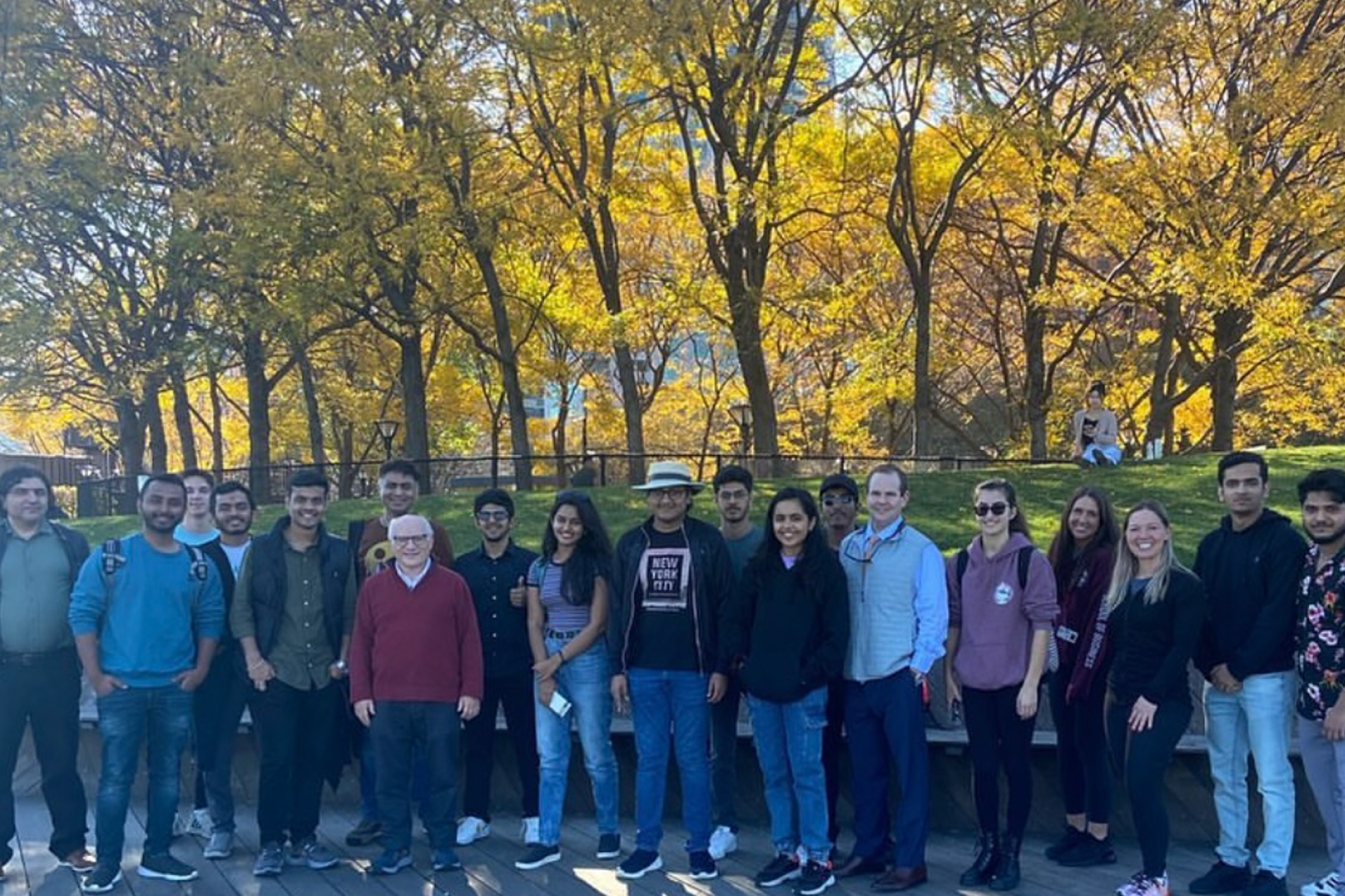 A group of students, faculty and staff pose for a group photo on a fall day with the sun shining through a group of yellow leafed trees.