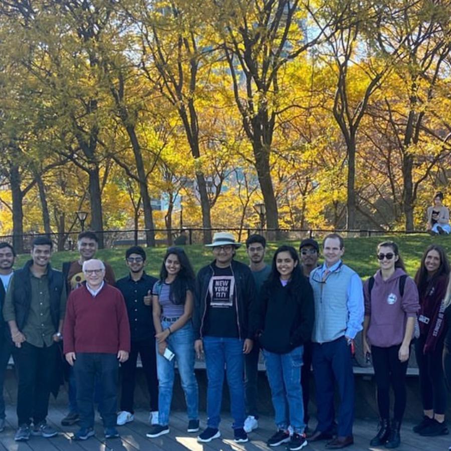 A group of students, faculty and staff pose for a group photo on a fall day with the sun shining through a group of yellow leafed trees.