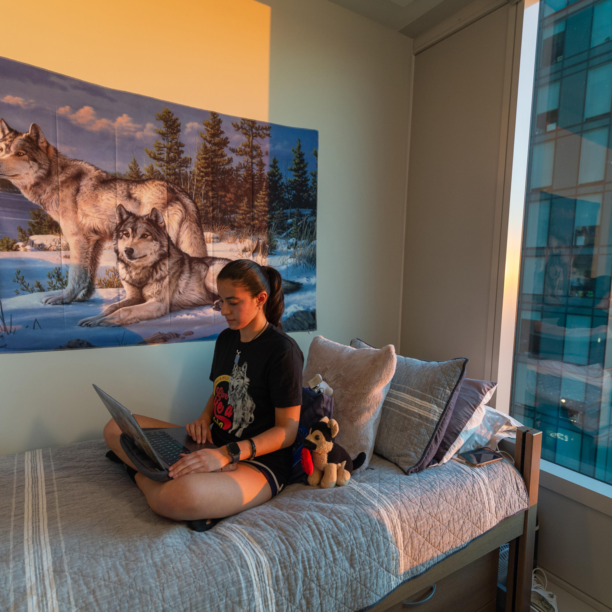 Student sits on her bed looking at her laptop with large wolf tapestry behind her.