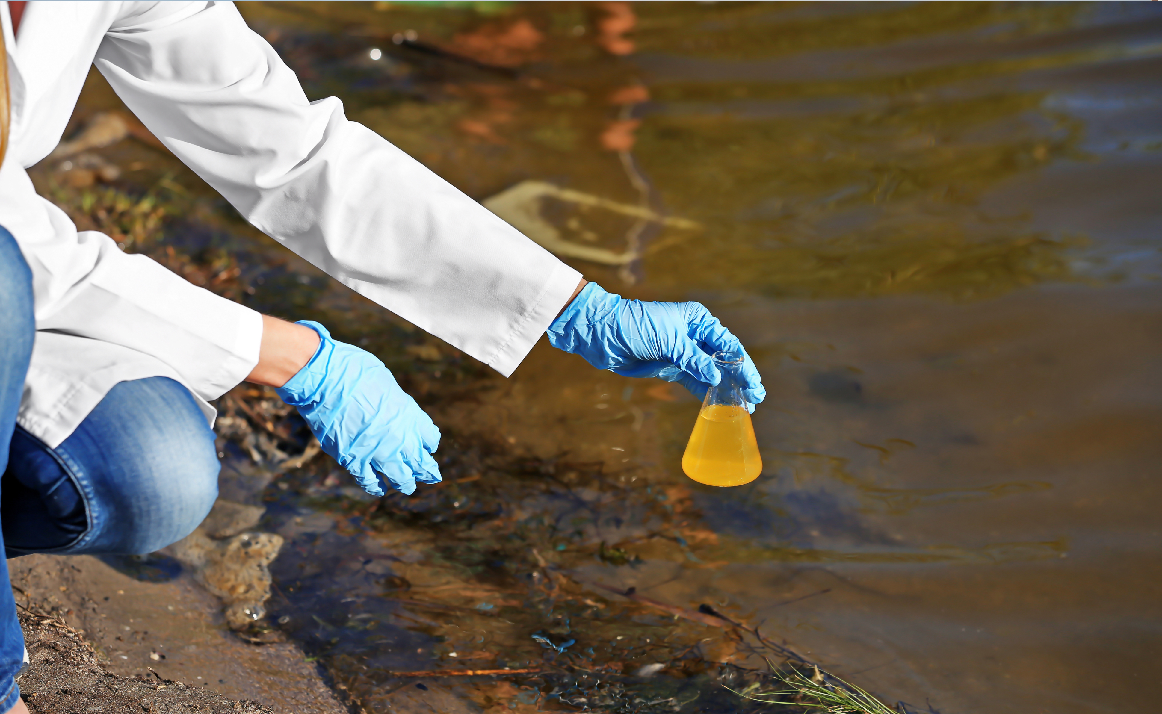 Unidentified person in lab coat and gloves holds beaker above body of water