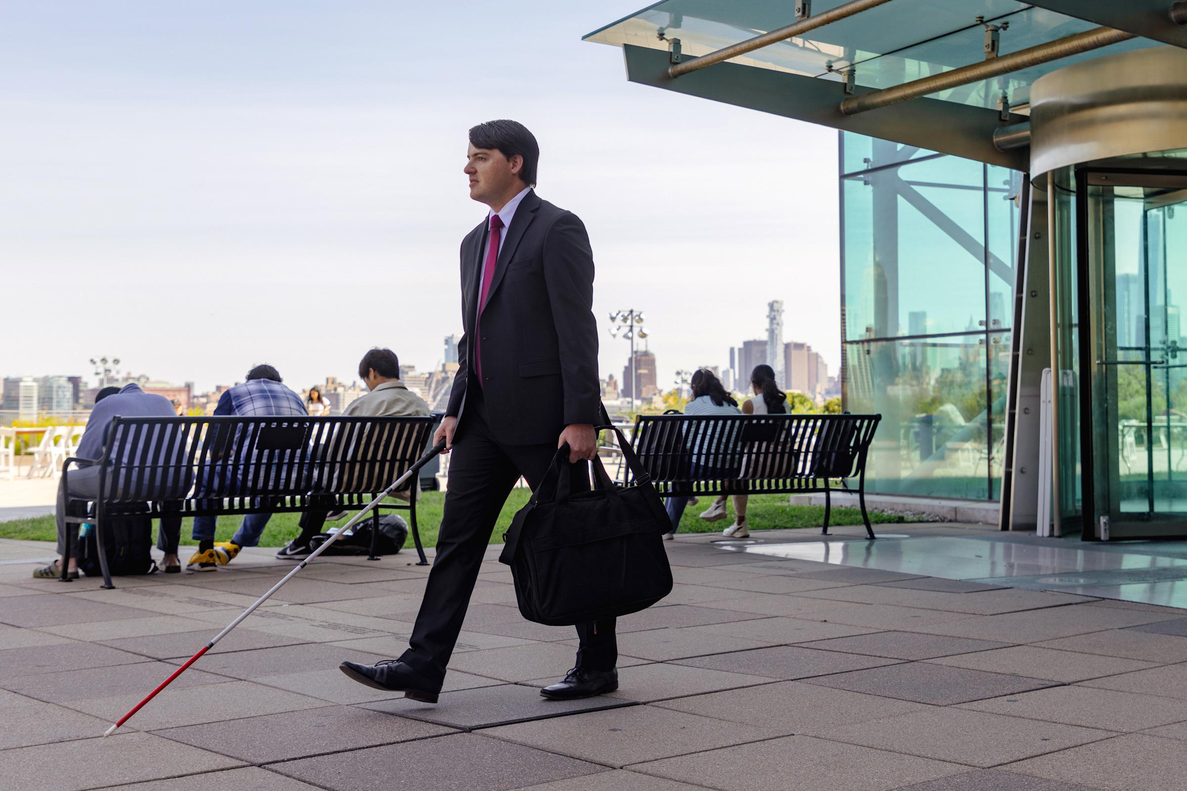 Robby Dahill walks with a cane outside the Babbio Center with NYC skyline in background.