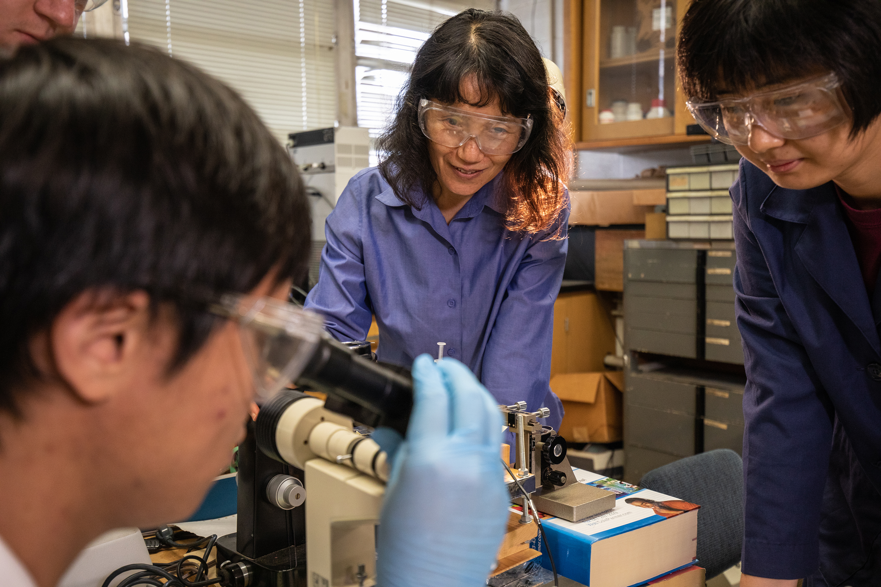 Hong Liang wears safety goggles and observes the students' work with a microscope