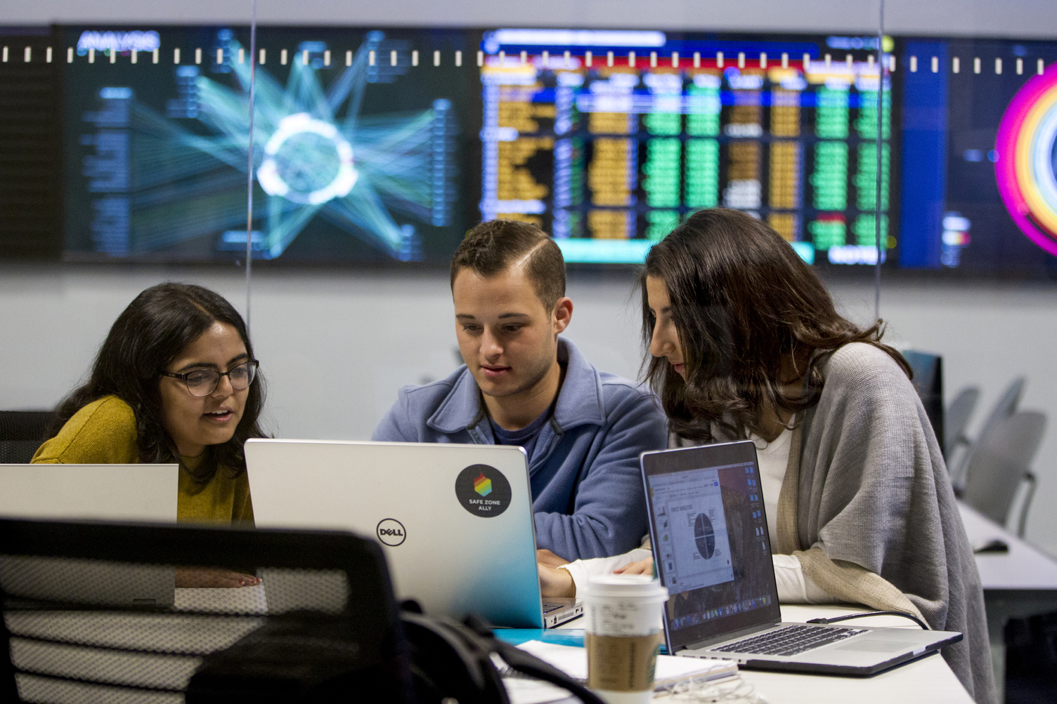 Three students looking at a laptop with a bank of monitors on a wall behind them.