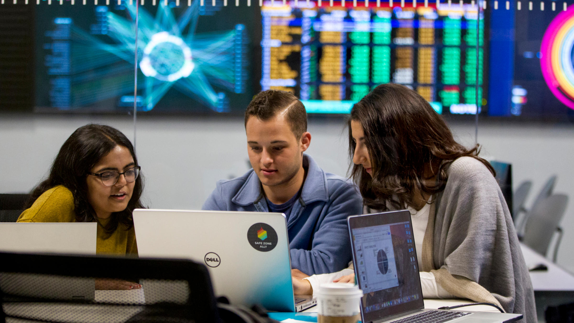 Three students looking at a laptop with a bank of monitors on a wall behind them.