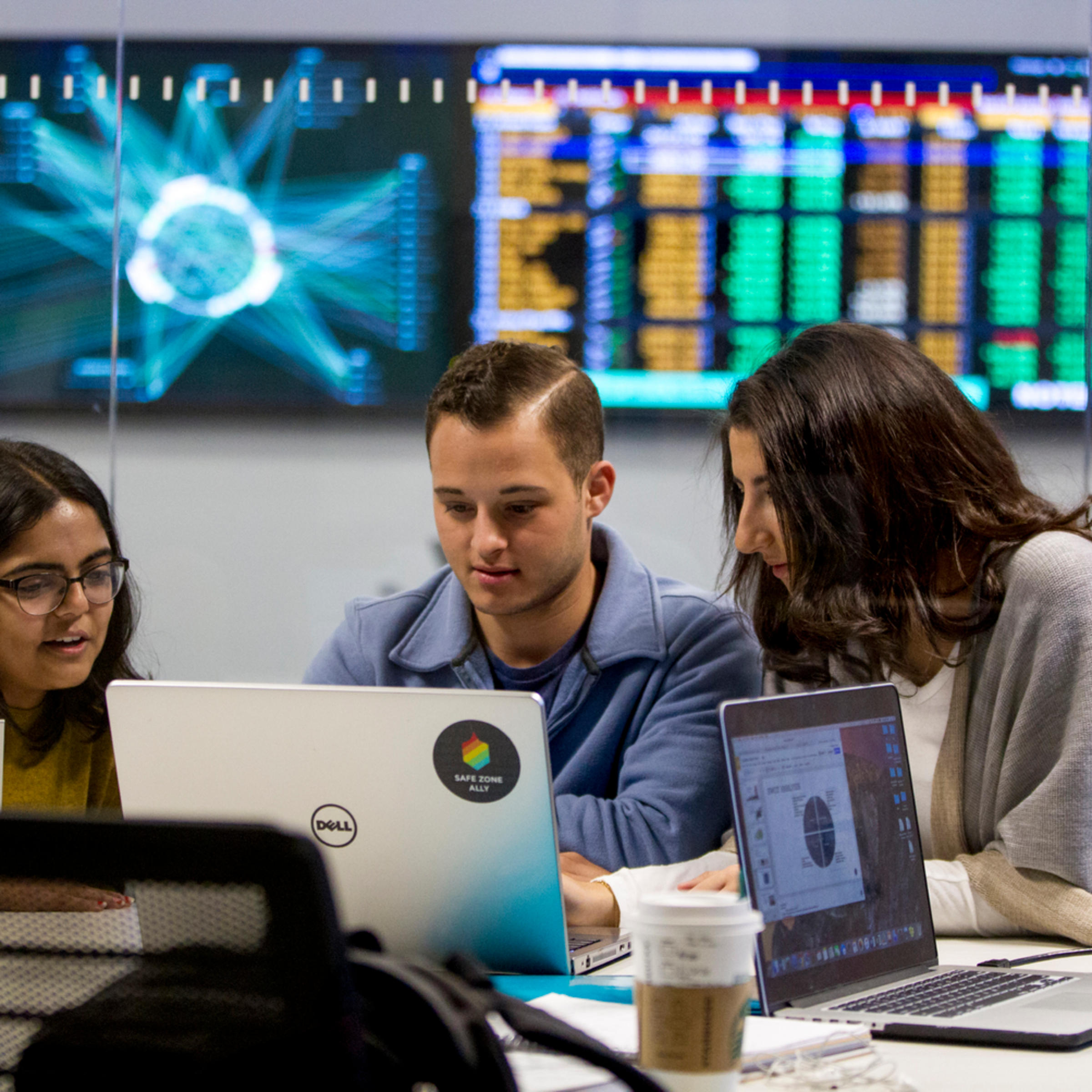 Three students looking at a laptop with a bank of monitors on a wall behind them.