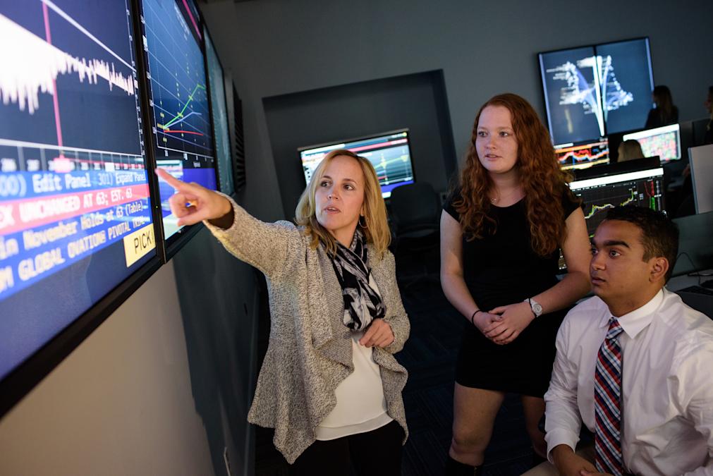 Instructor points to data on large wall monitor white two students look on, interested