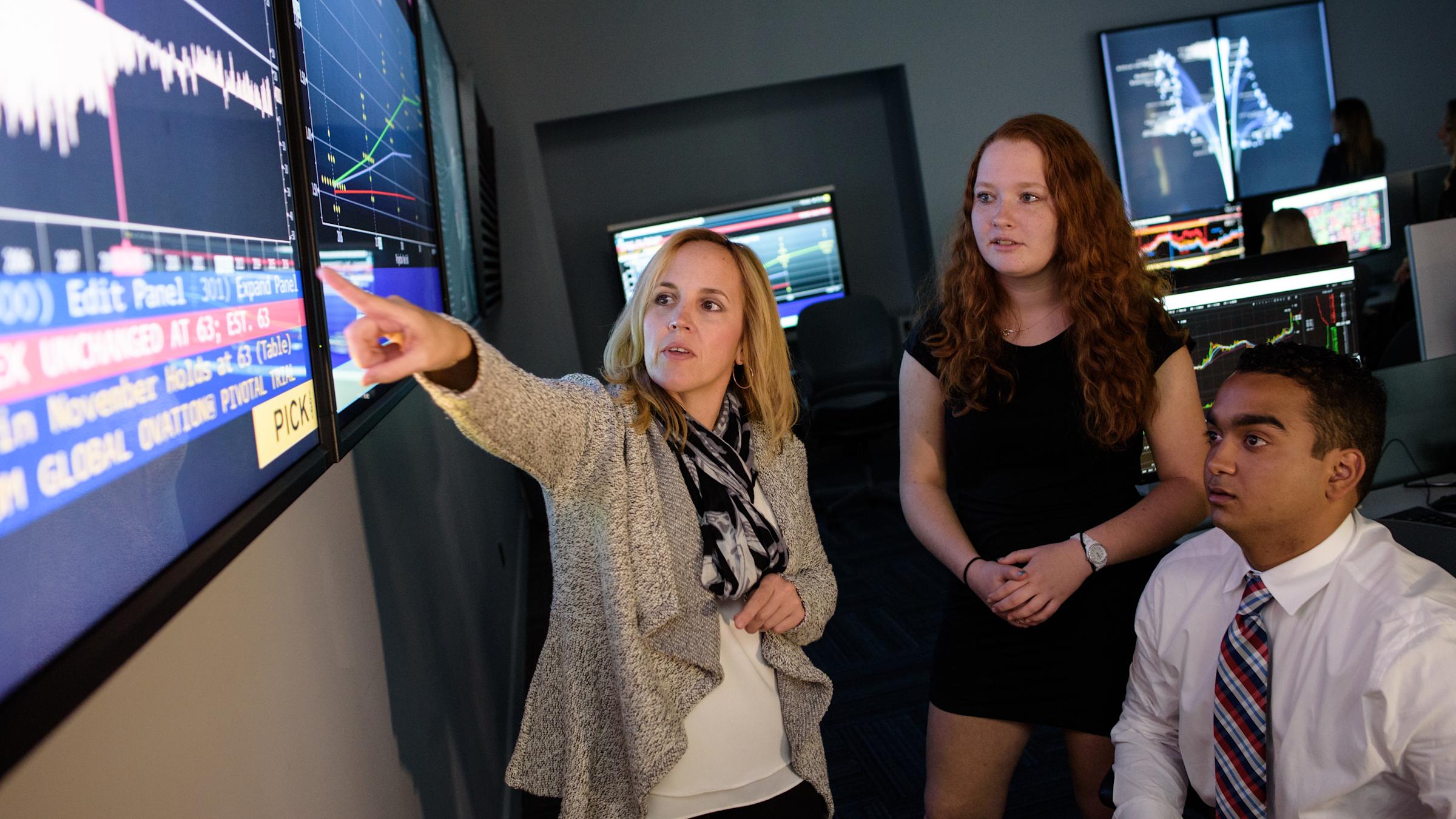 Instructor points to data on large wall monitor white two students look on, interested