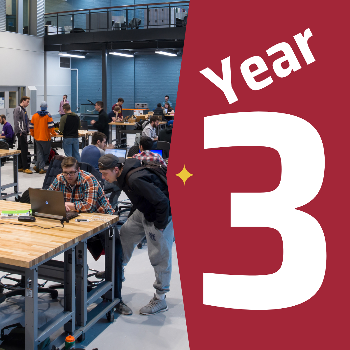 Two students standing in the midst of a crowd in the ABS Engineering Center working on a laptop at one of the tables next to a graphic that says year three.