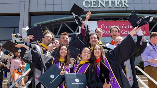 Students pose and smile in cap and gown, holding their diplomas, on the steps of Meadowlands Exposition Center