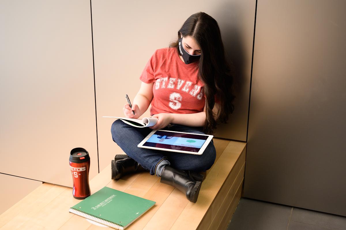 Student with green notebook and red water bottle taking notes and reading off iPad