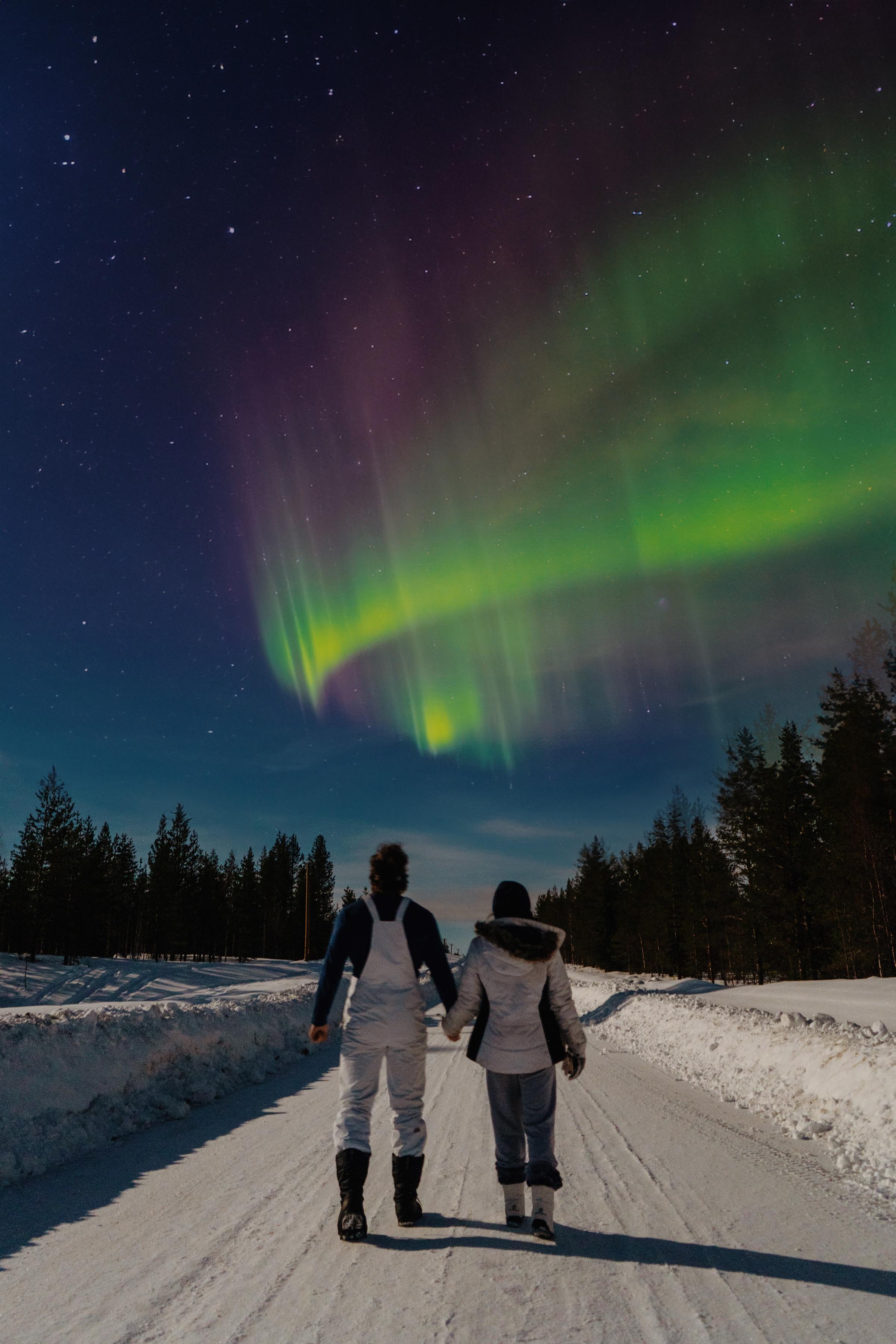 Two people in winter gear hold hands while walking down a snow-covered road toward a breathtaking aurora borealis display of green, pink, and purple lights swirling across a starry night sky, framed by dark silhouetted pine trees.