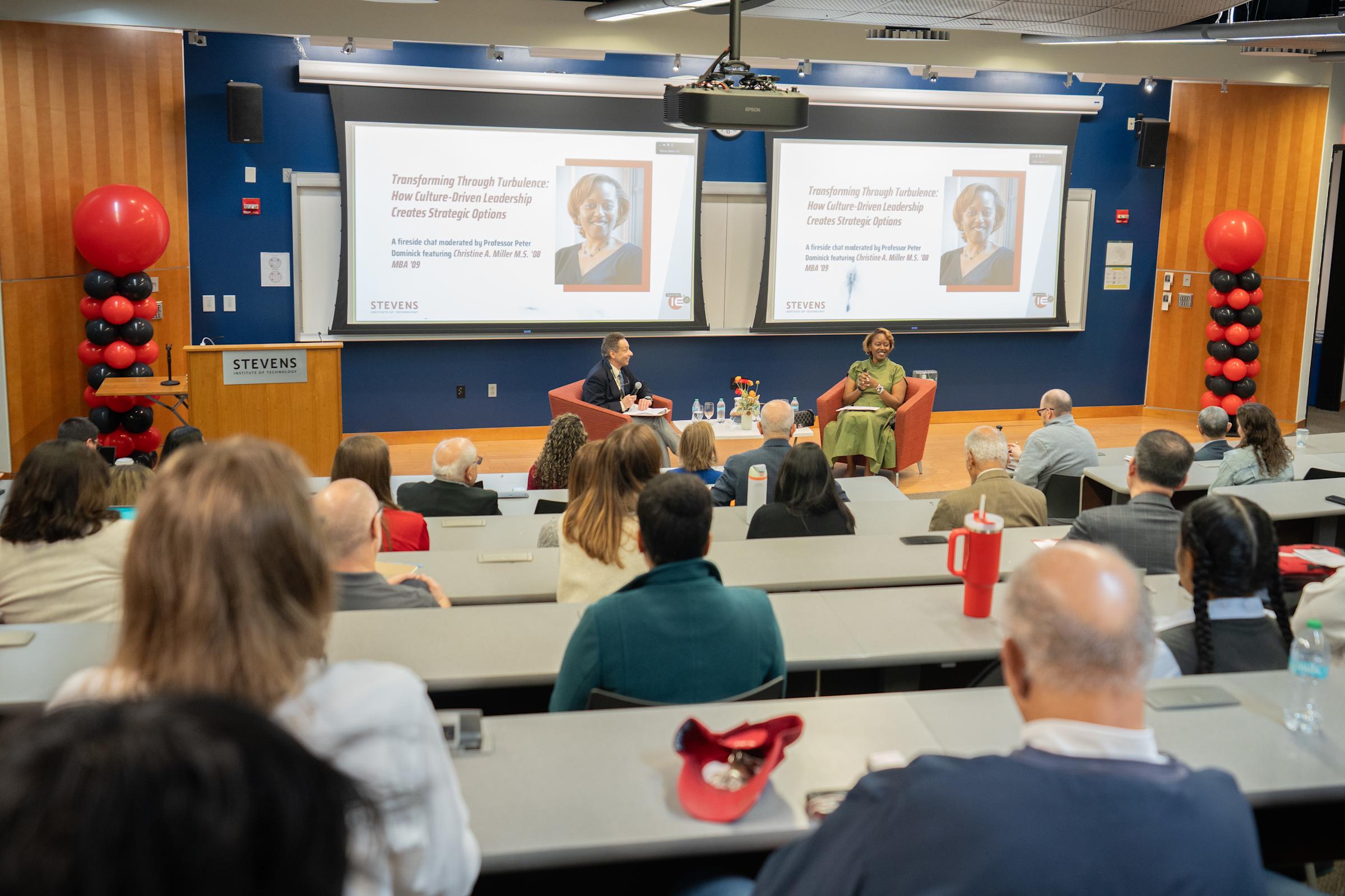 Fireside chat with two speakers in orange chairs on stage. Dual screens display presentation on culture-driven leadership. Audience seated at tiered tables in modern lecture hall with red/black balloon décor and blue walls.
