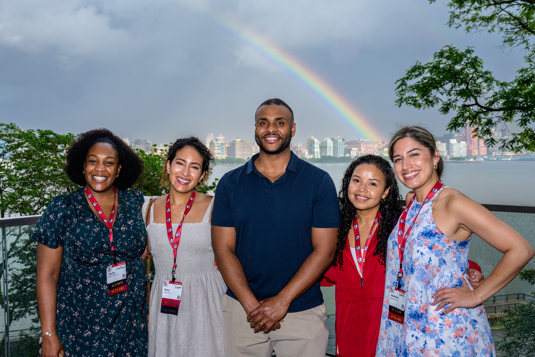 Five young alumni smile and pose at Babbio patio in front of a rainbow during Alumni Weekend 2025