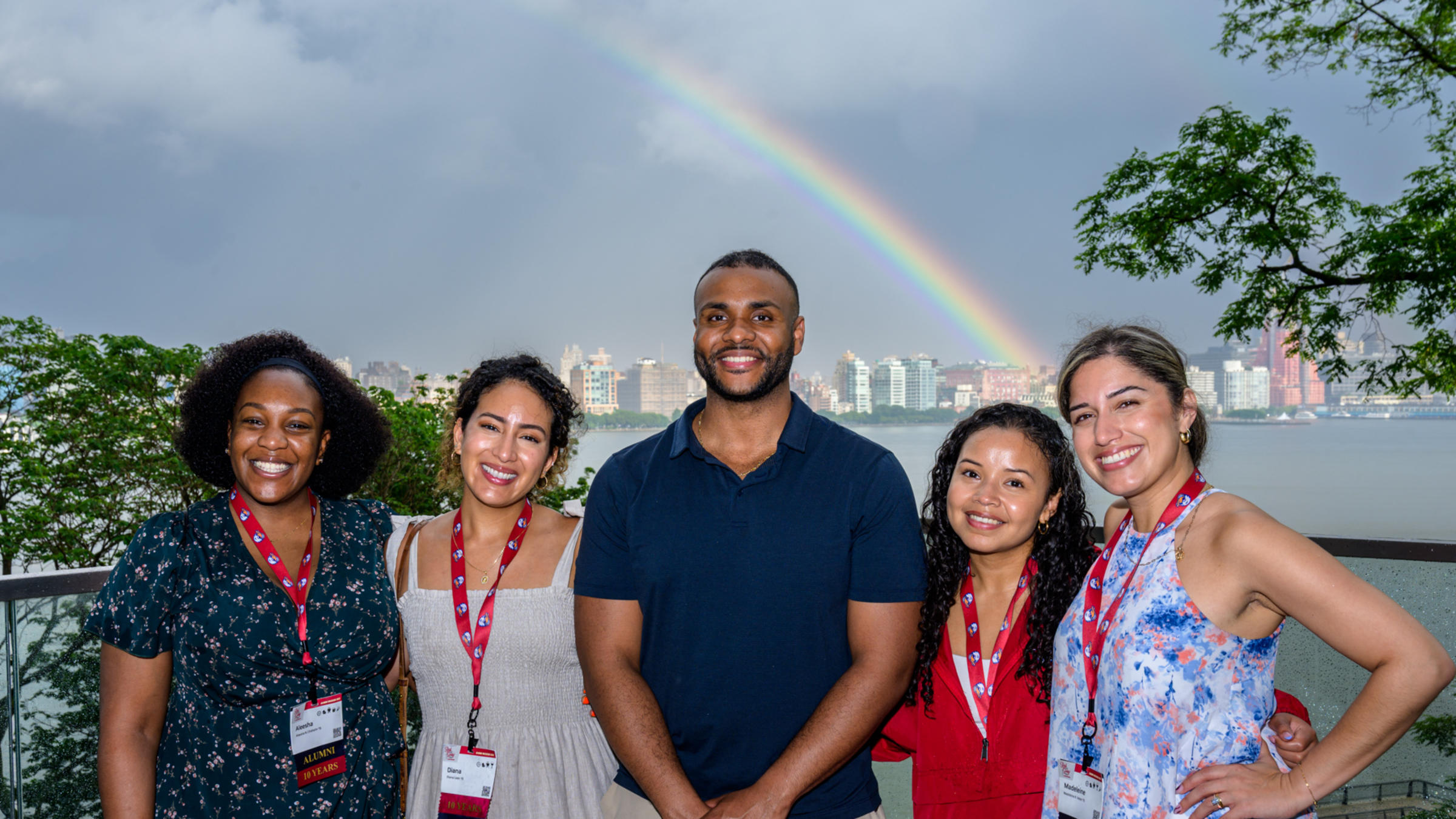 Five young alumni smile and pose at Babbio patio in front of a rainbow during Alumni Weekend 2025