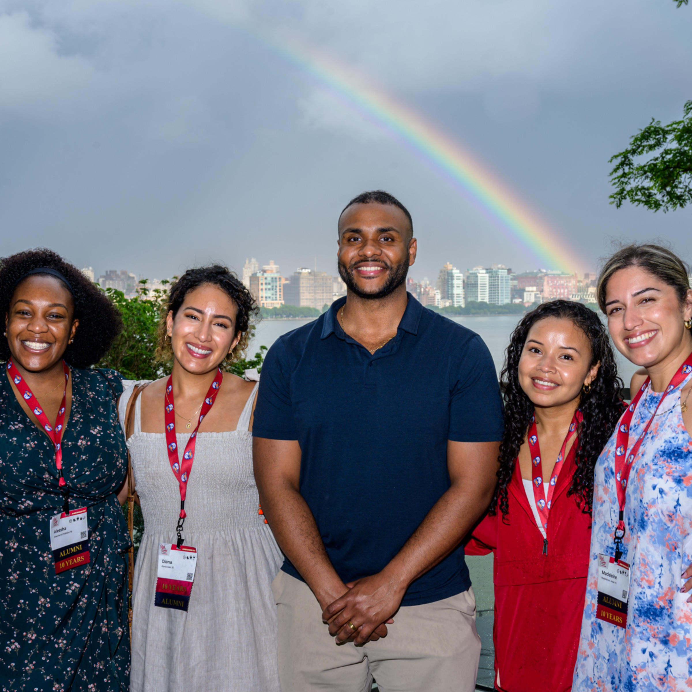 Five young alumni smile and pose at Babbio patio in front of a rainbow during Alumni Weekend 2025