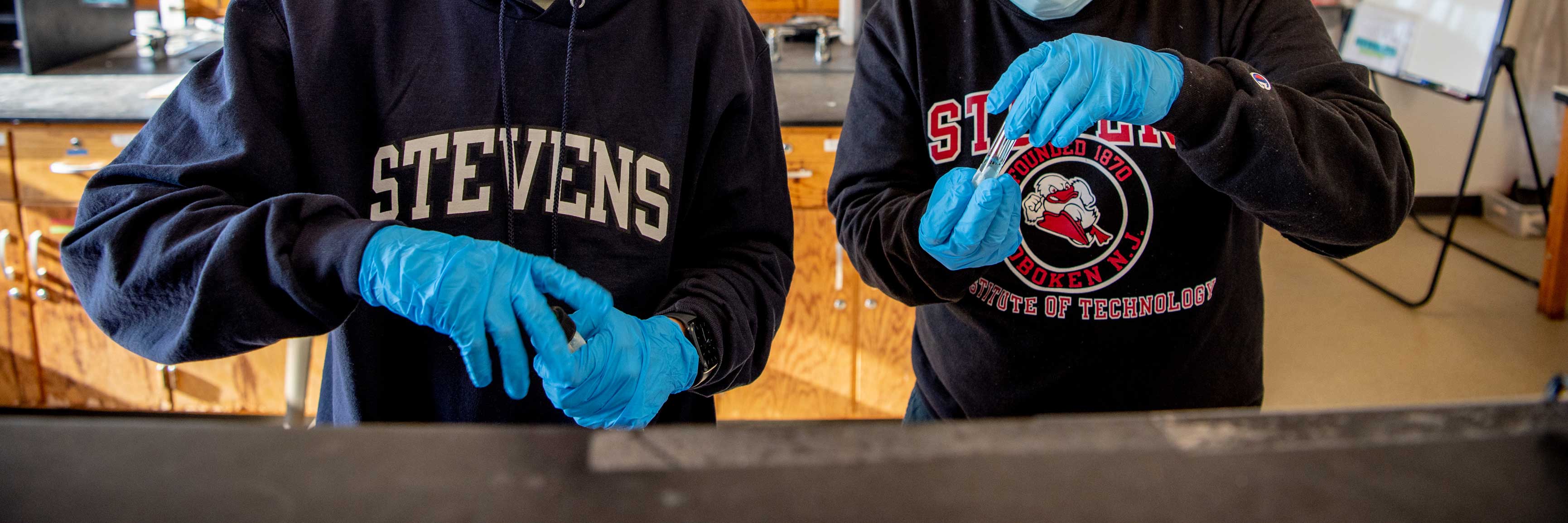Two pairs of students gloved hands work with chemistry equipment while wearing Stevens apparel.