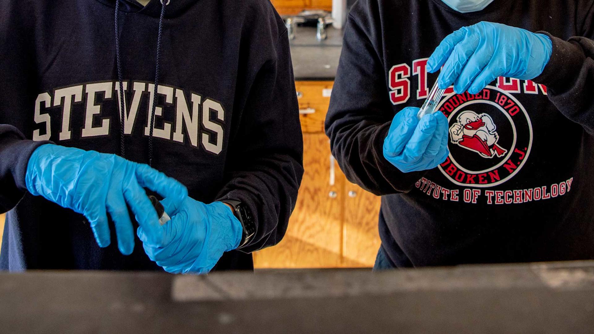 Two pairs of students gloved hands work with chemistry equipment while wearing Stevens apparel.