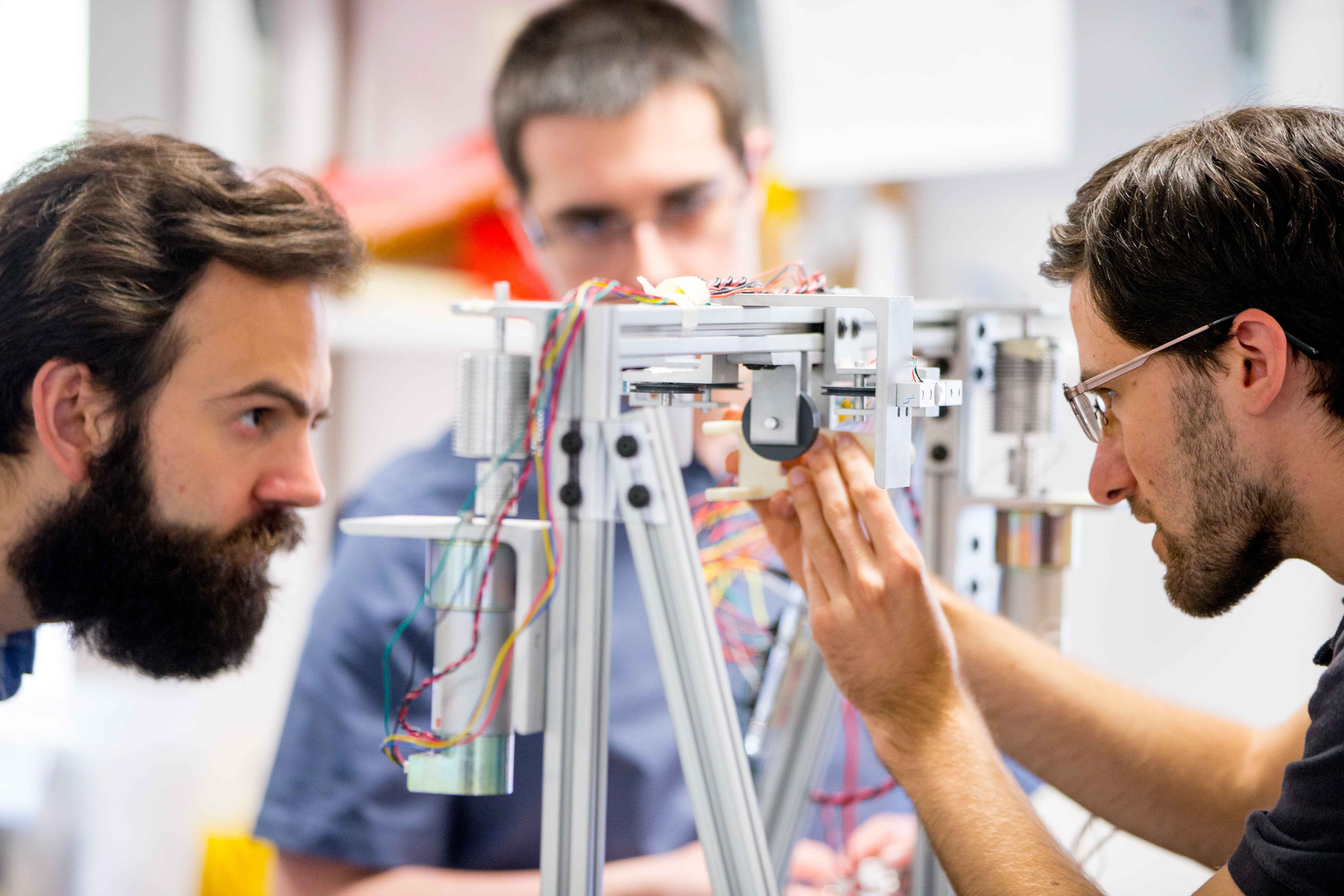 Three male student engineers working on a prototype made of metal piping and wires.