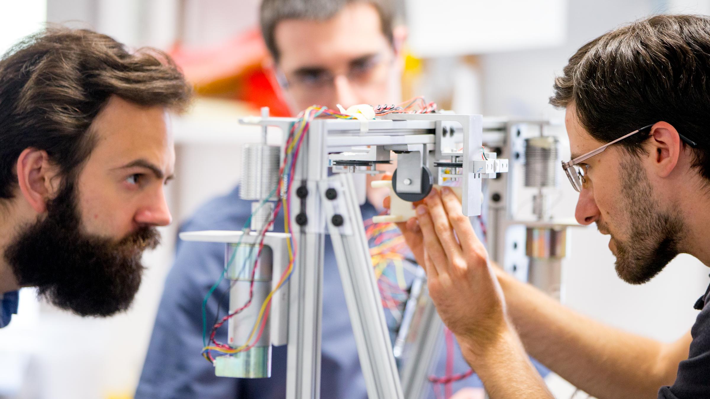 Three male student engineers working on a prototype made of metal piping and wires.