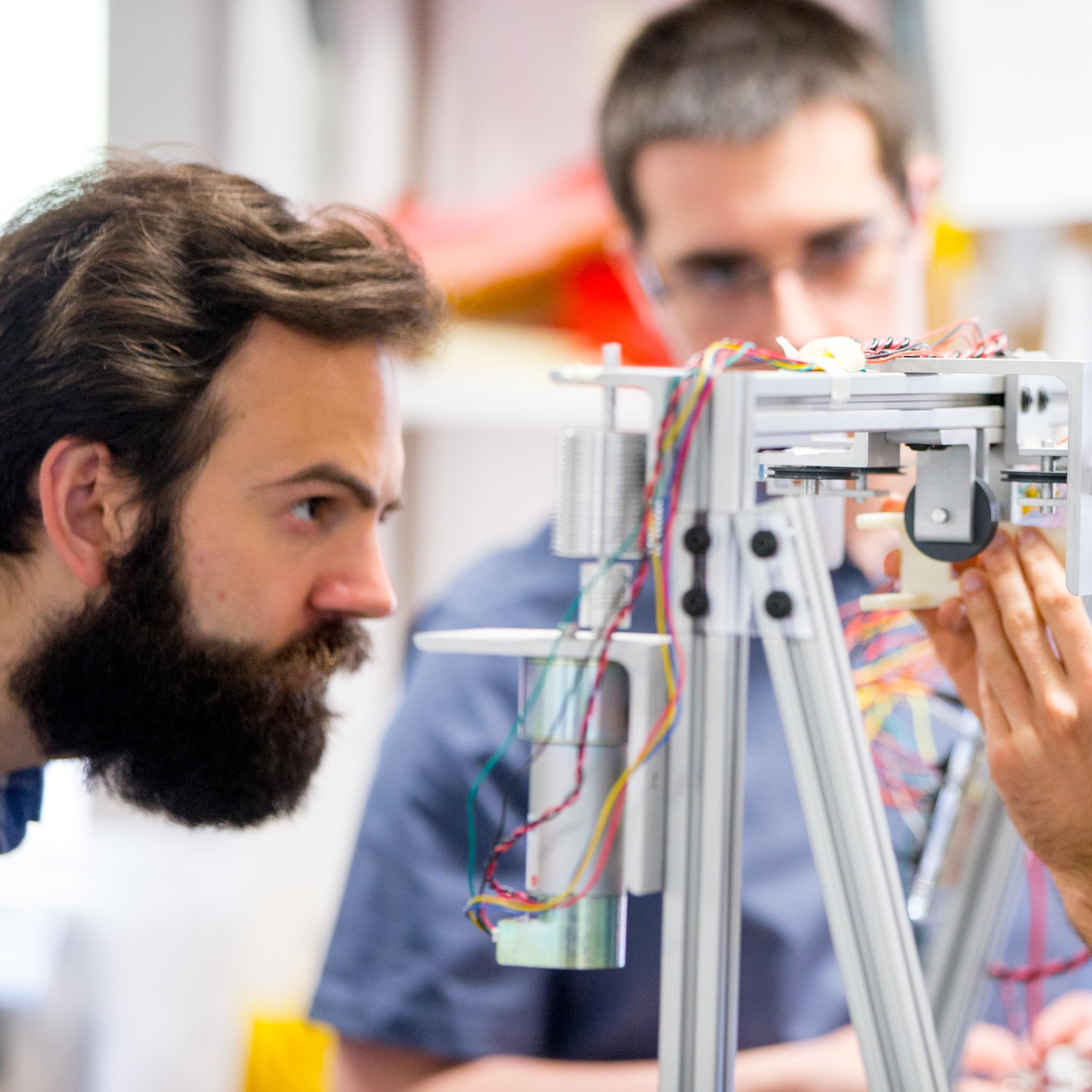 Three male student engineers working on a prototype made of metal piping and wires.