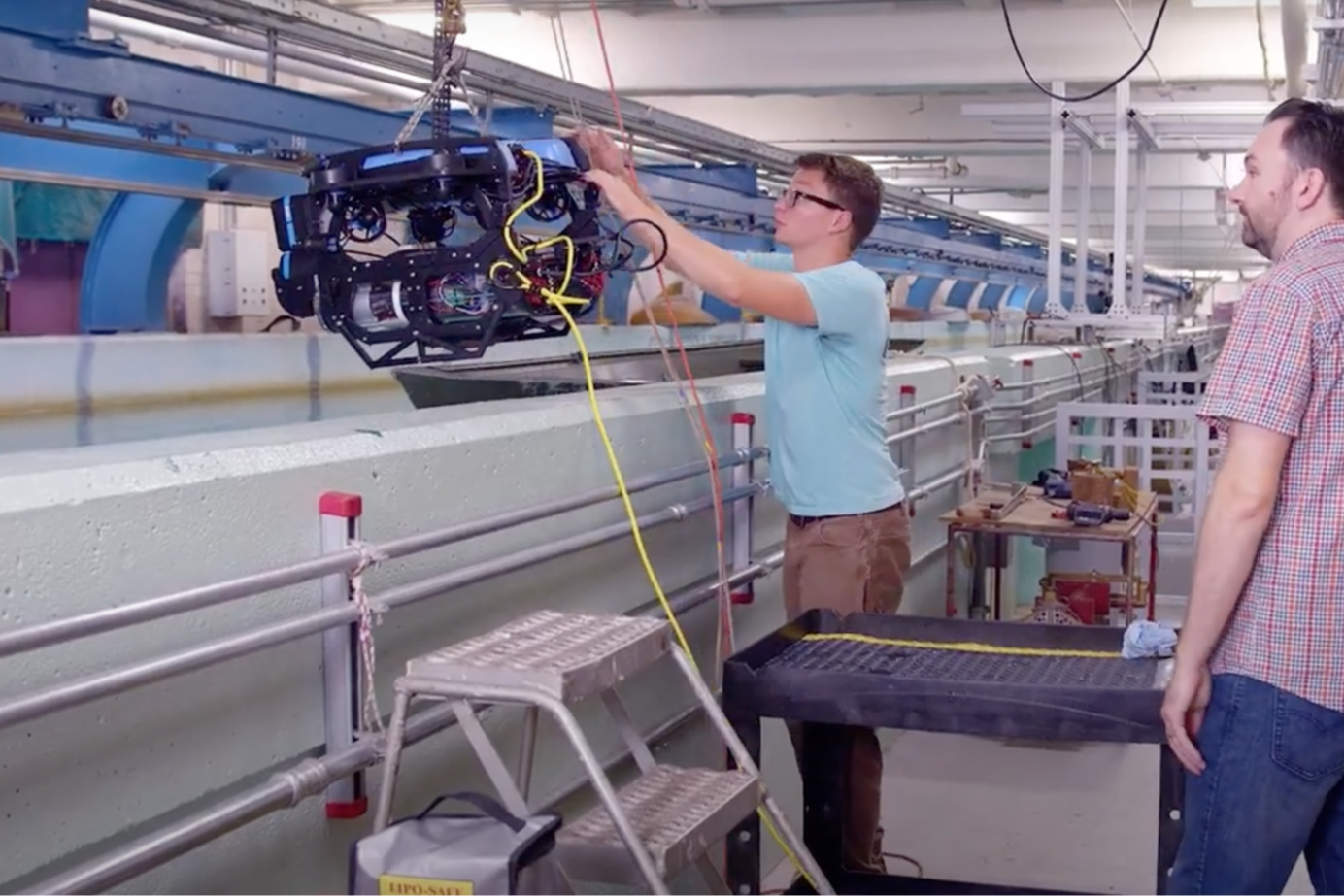 A student moves an underwater robot into a testing water tank while a professor and another student assist.