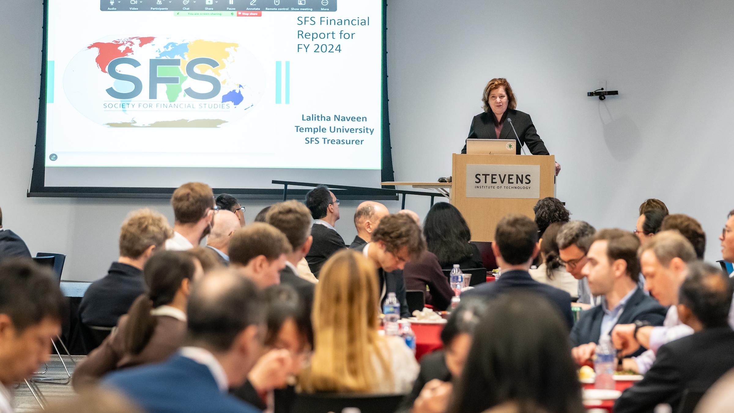A woman stands on stage behind a podium in front of a slide with the SFS Cavalcade logo.