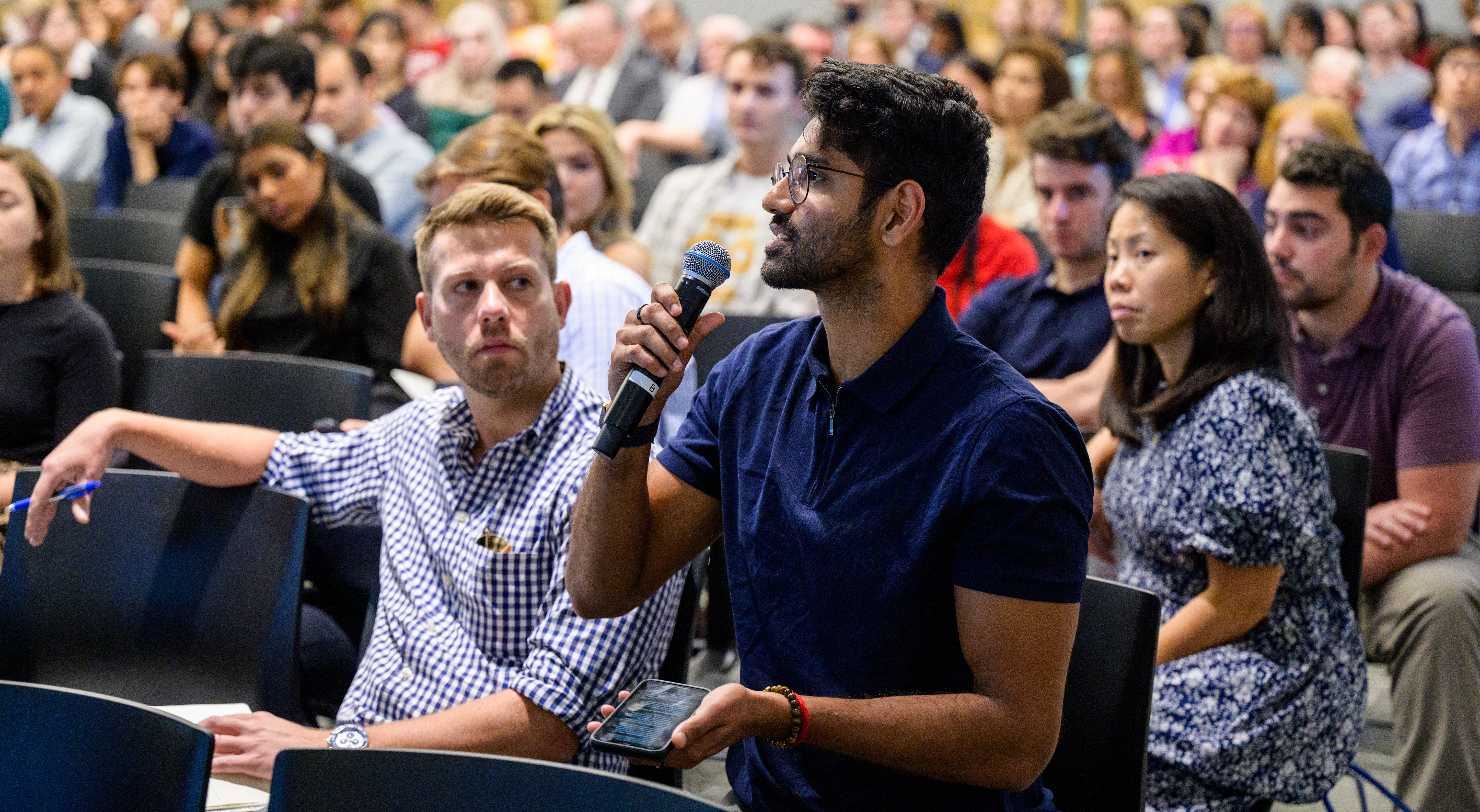 A man in a blue shirt speaks into a microphone during a seminar, seated among a large audience. 