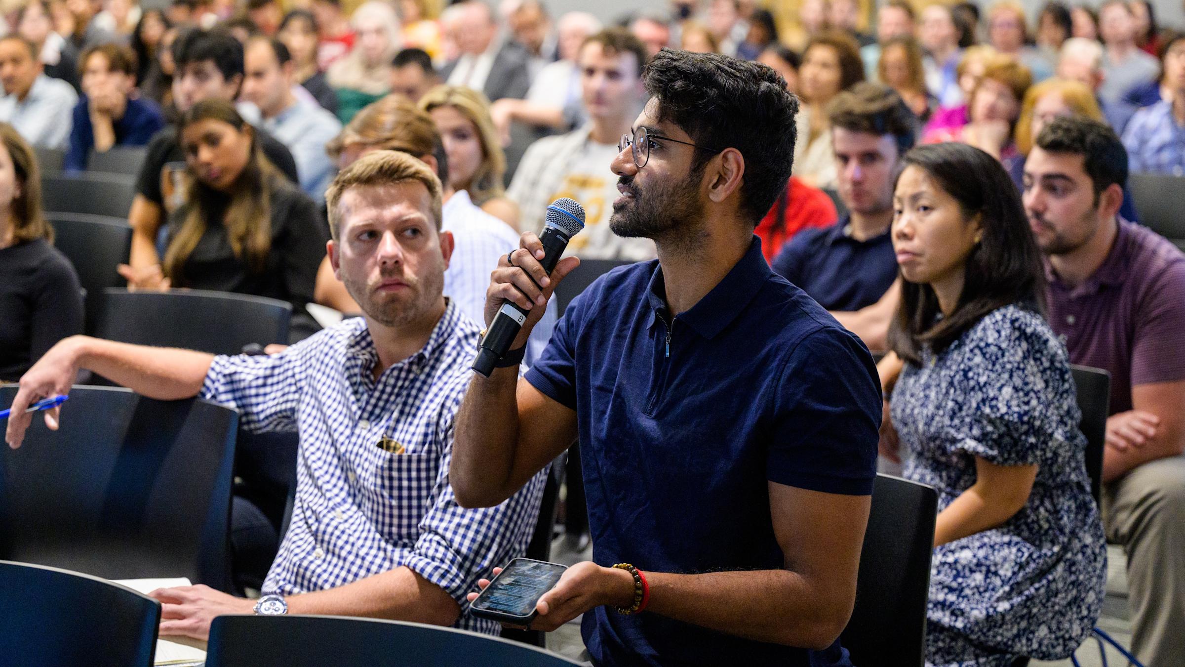 A man in a blue shirt speaks into a microphone during a seminar, seated among a large audience.