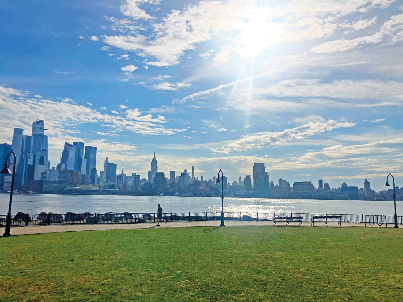 Photo of Midtown Manhattan, the Hudson River and Maxwell Place Park, taken from the Hoboken waterfront, with a blue sky, sun peeking from the clouds and a lone person jogging.
