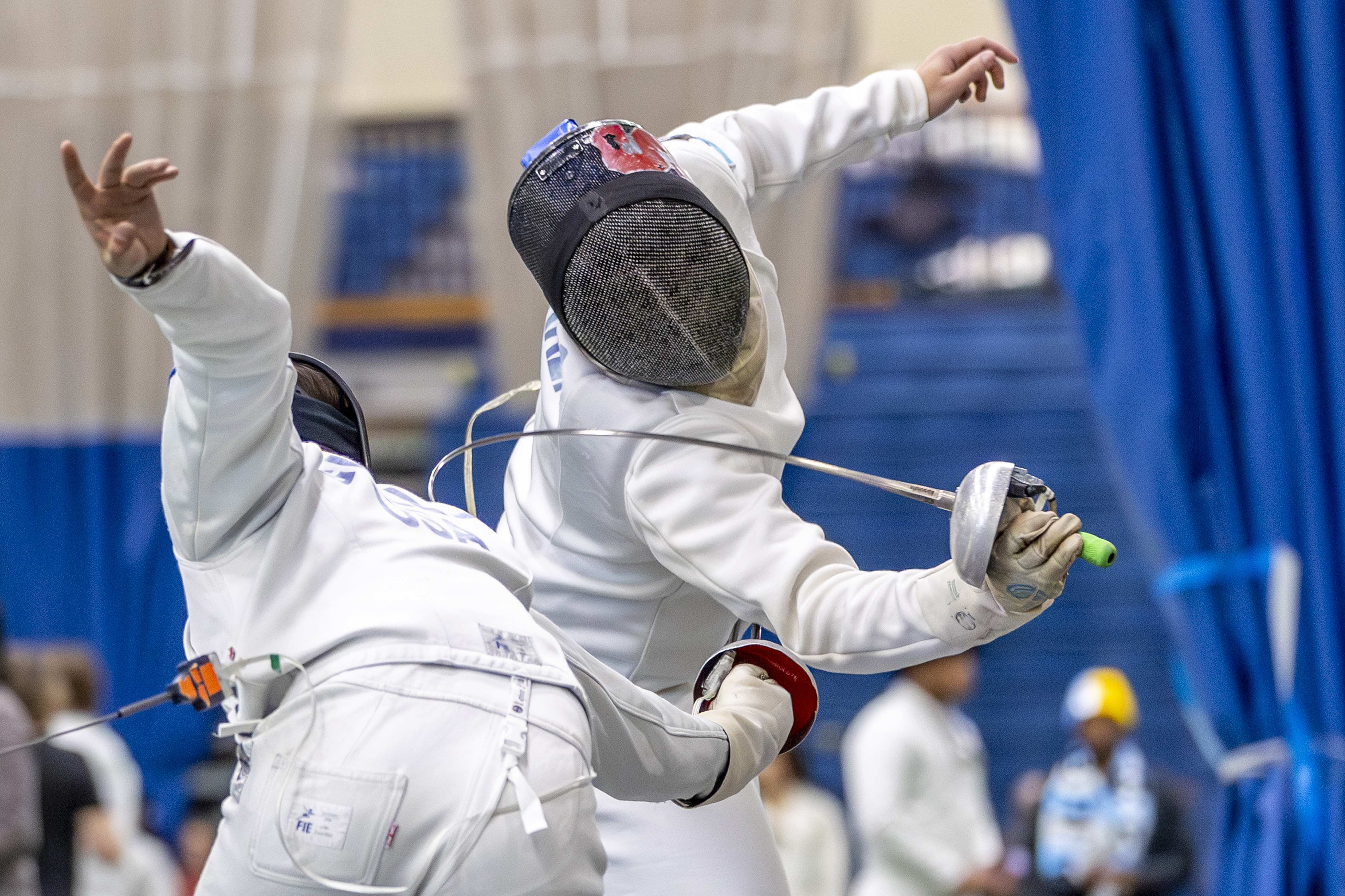 Two fencers in full protective gear compete during a bout. One fencer lunges forward with an épée extended while the other leans back to evade.