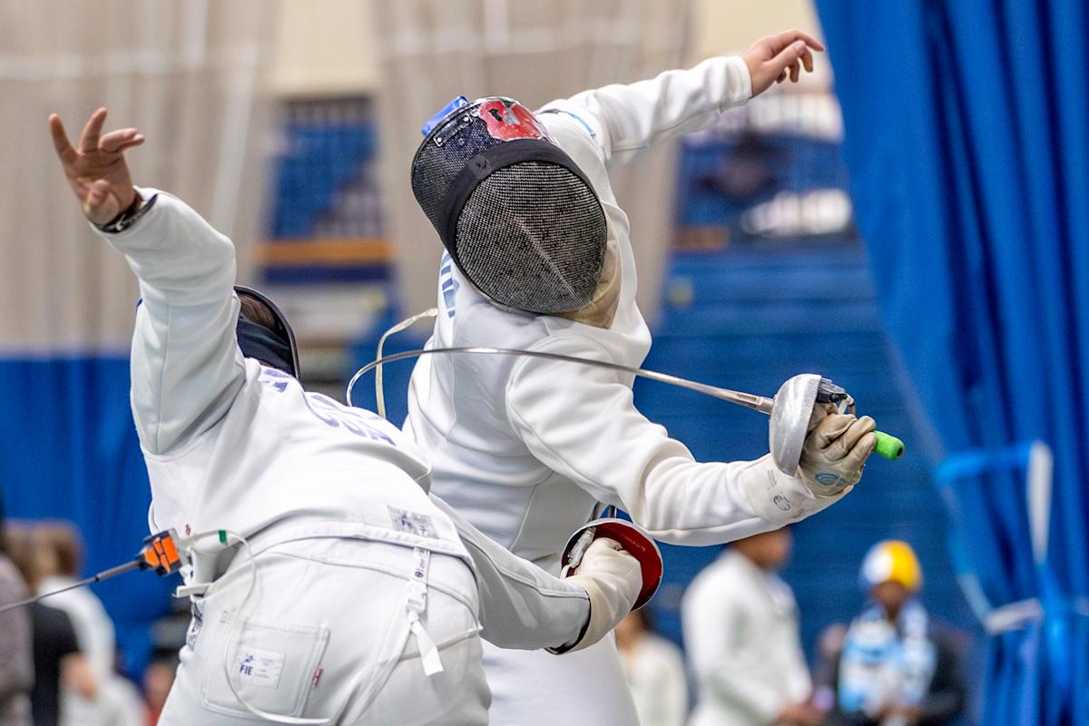 Two fencers in full protective gear compete during a bout. One fencer lunges forward with an épée extended while the other leans back to evade.