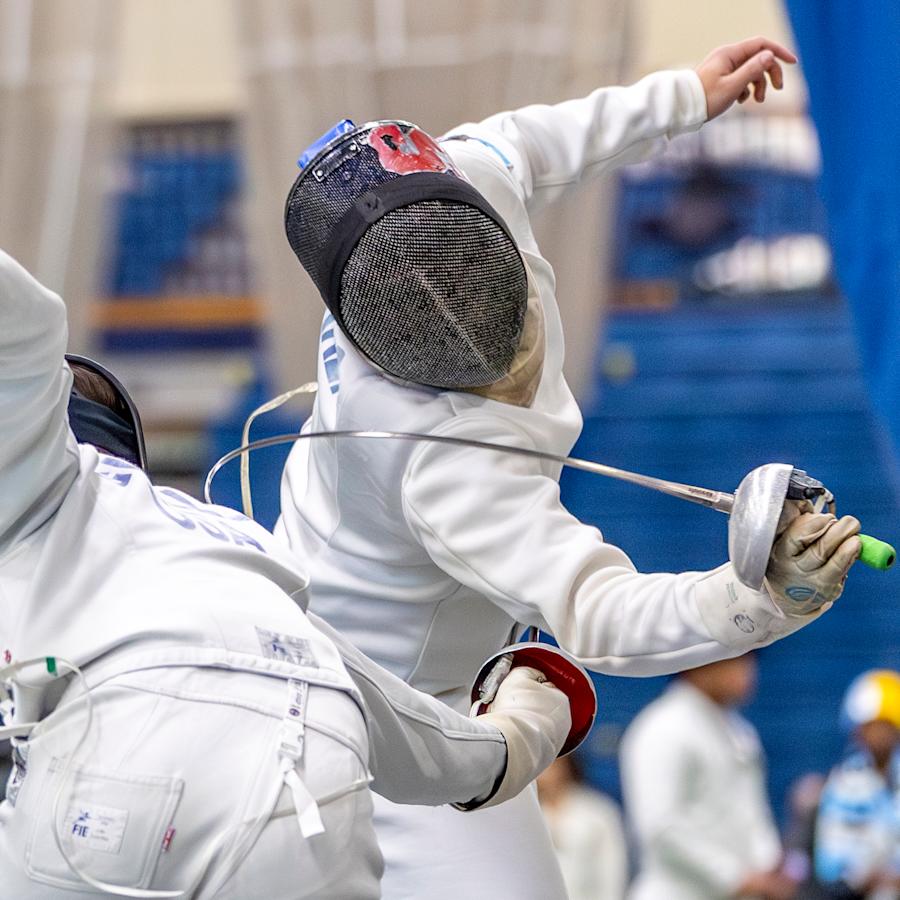 Two fencers in full protective gear compete during a bout. One fencer lunges forward with an épée extended while the other leans back to evade.