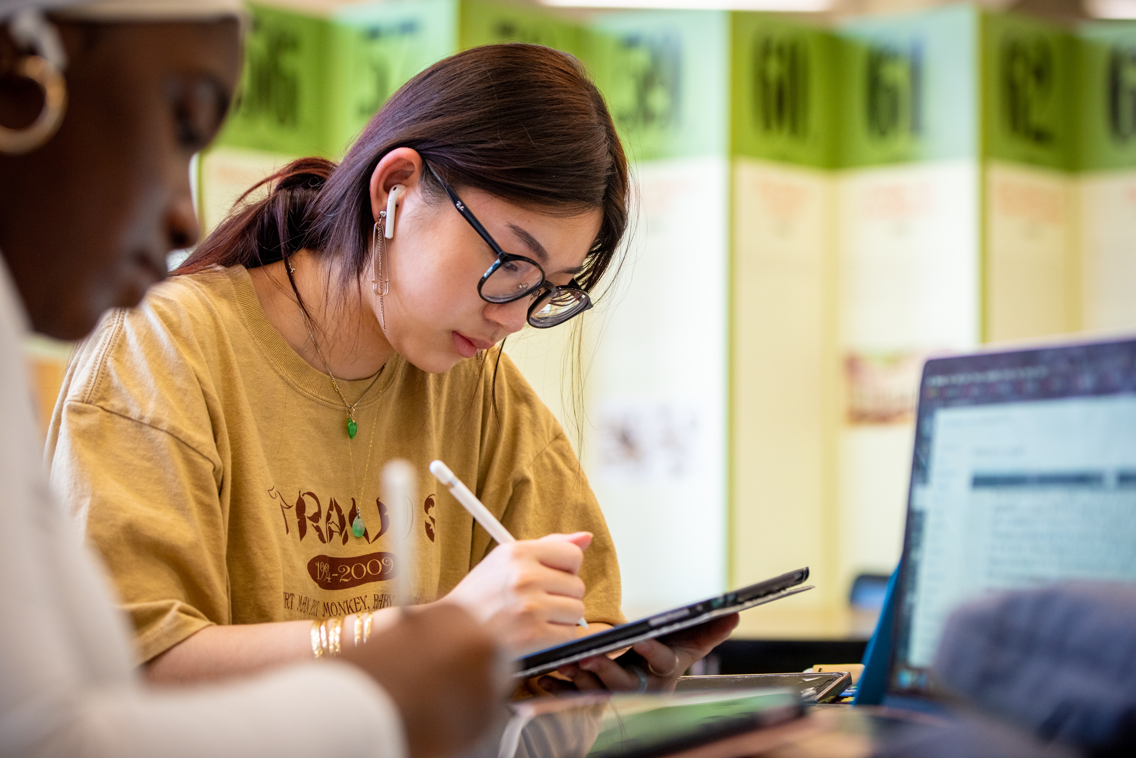 Female student writes on tablet in classroom. Another female student also works on a computer.