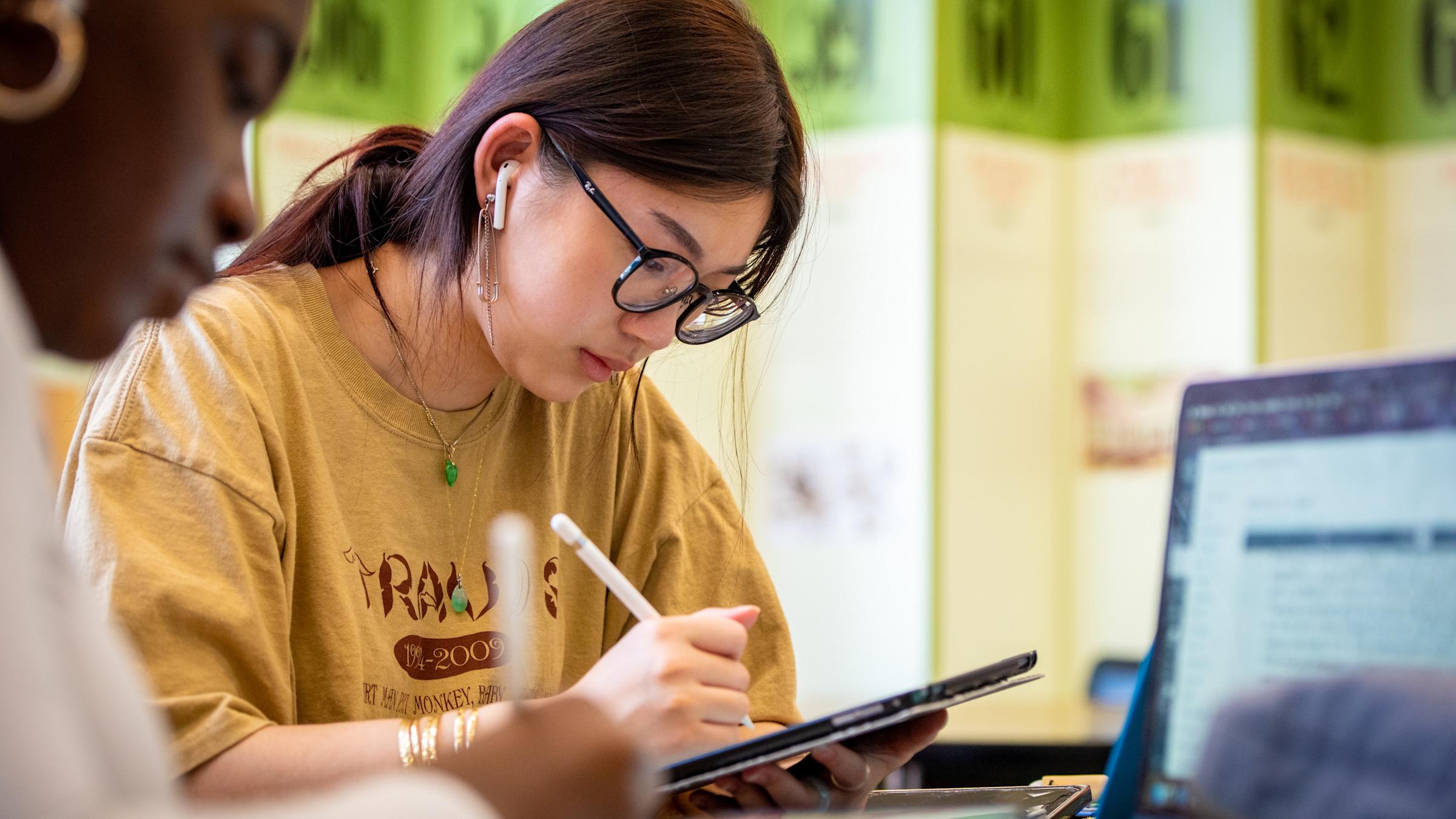 Female student writes on tablet in classroom. Another female student also works on a computer.