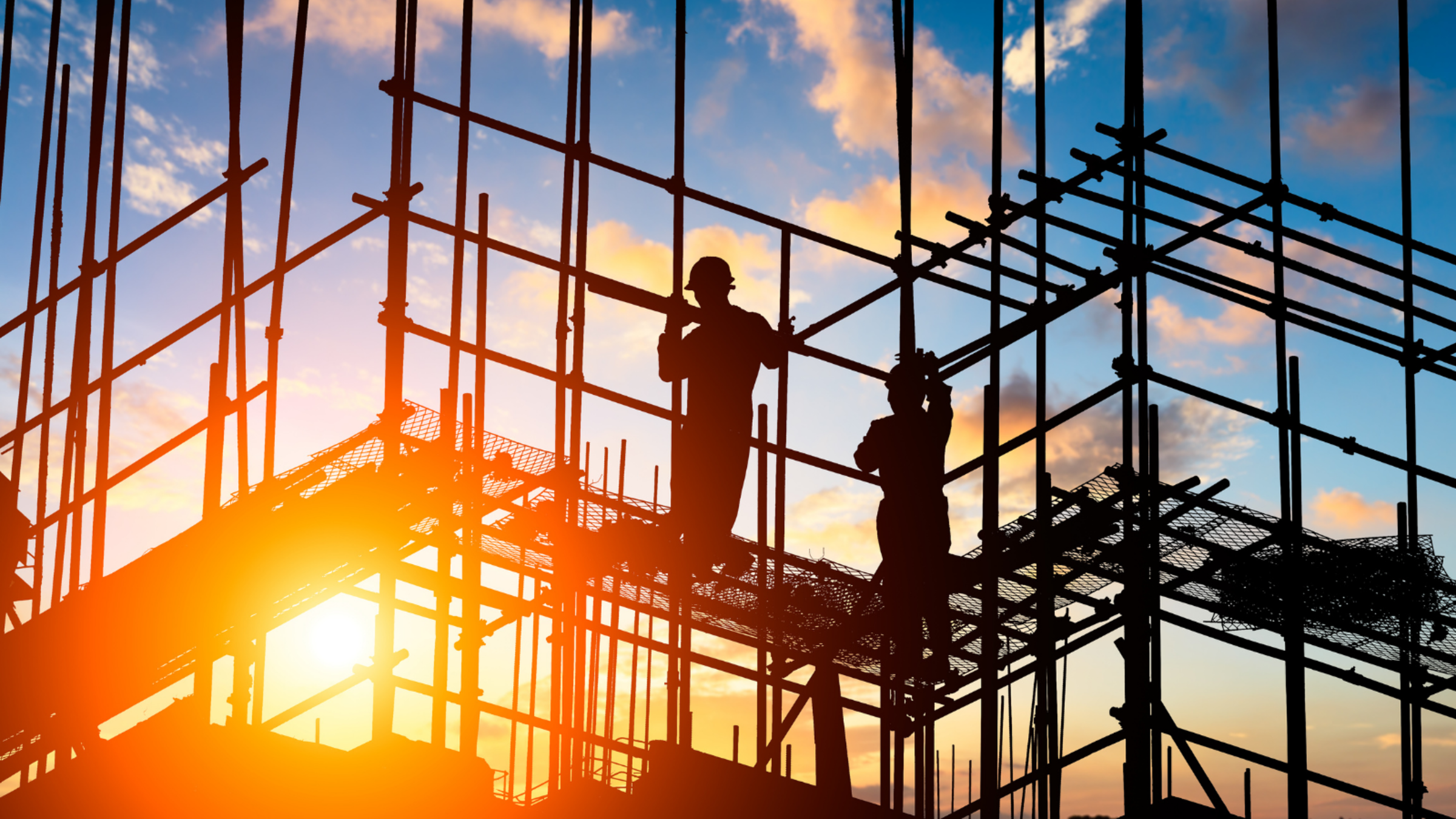 Silhouetted construction workers framed by steel scaffolding and rebar on a building site as the sun sets behind them with a warm orange glow and clouds in the sky.