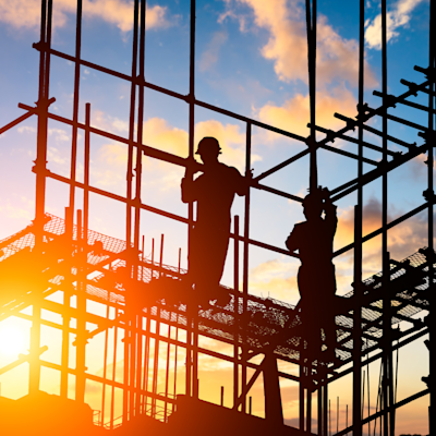 Silhouetted construction workers framed by steel scaffolding and rebar on a building site as the sun sets behind them with a warm orange glow and clouds in the sky.