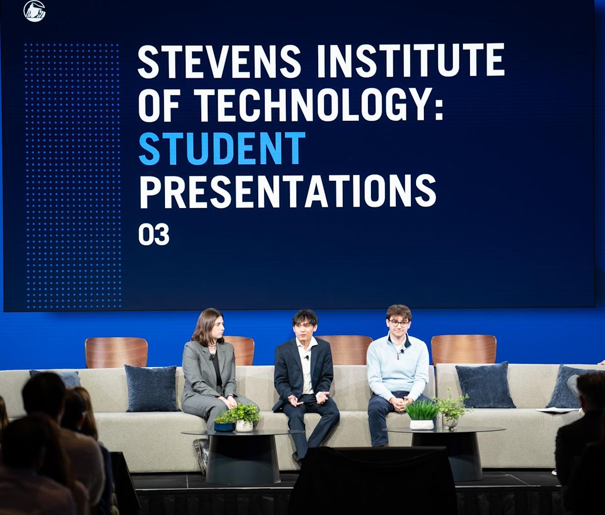 Three students sit on stage under a screen that reads "Stevens Institute of Technology: Student Presentations"