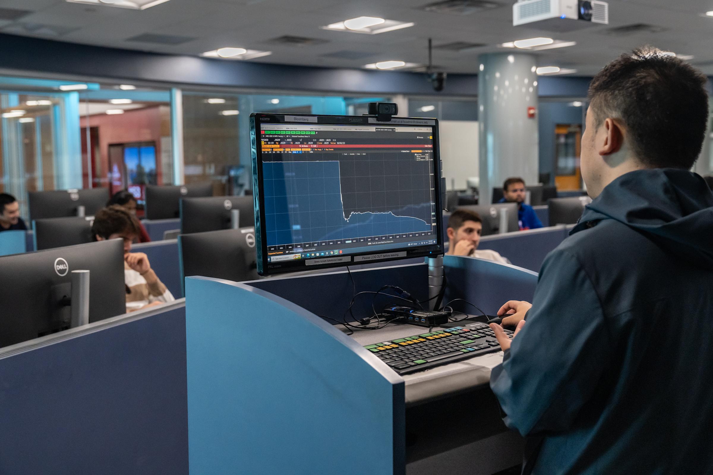 A student works at a trading terminal with specialized Bloomberg keyboard and monitor showing market data and price charts. Other students work at individual workstations in the modern financial lab with blue partitions and glass-walled offices visible.