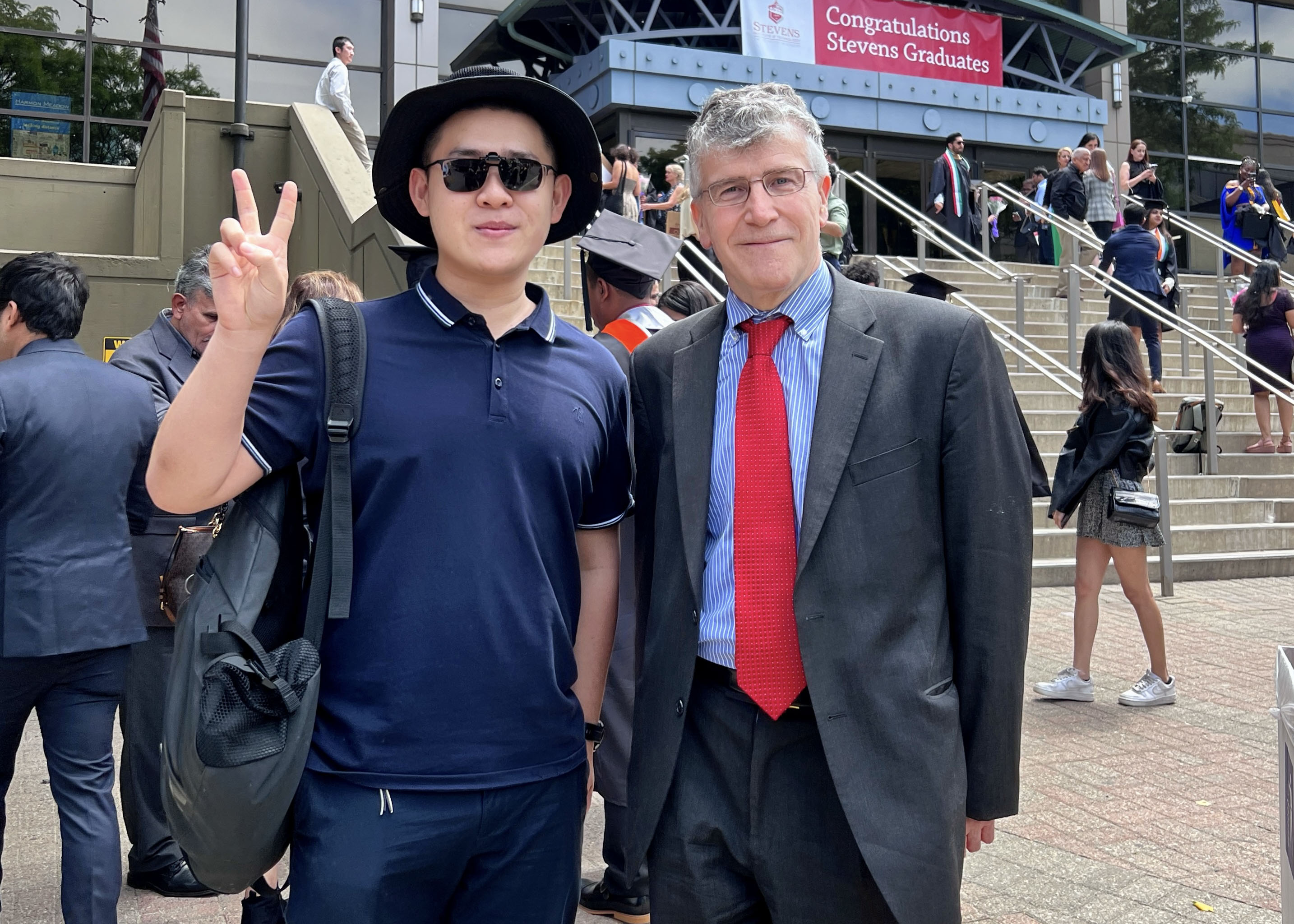 Rui Zong holds up two fingers standing next to professor Emmanuel Hatzakis.