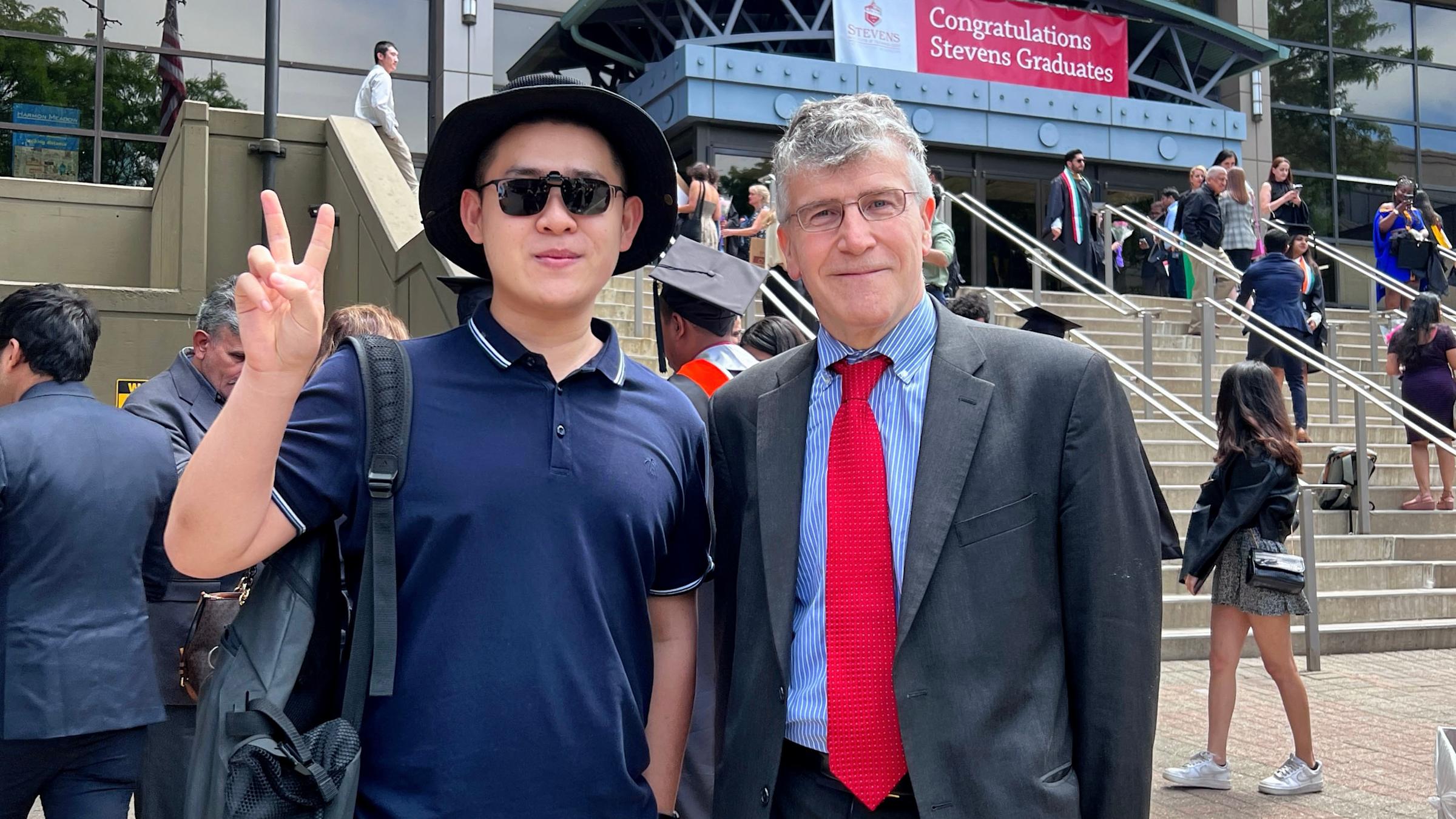 Rui Zong holds up two fingers standing next to professor Emmanuel Hatzakis.