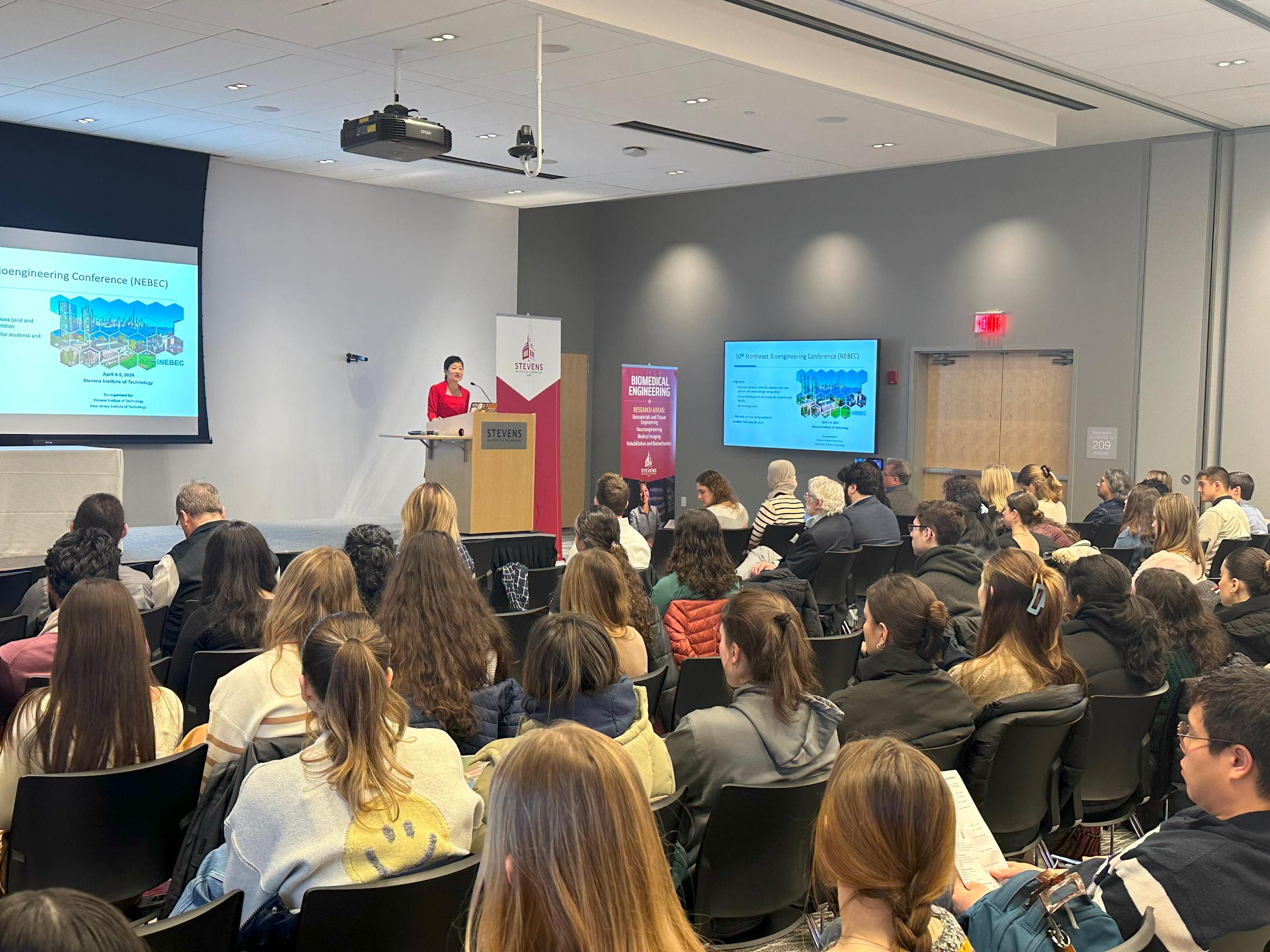 Jennifer Kang-Mieler stands in front of a crowd, behind a podium, with a projected image behind her, delivering welcoming remarks.