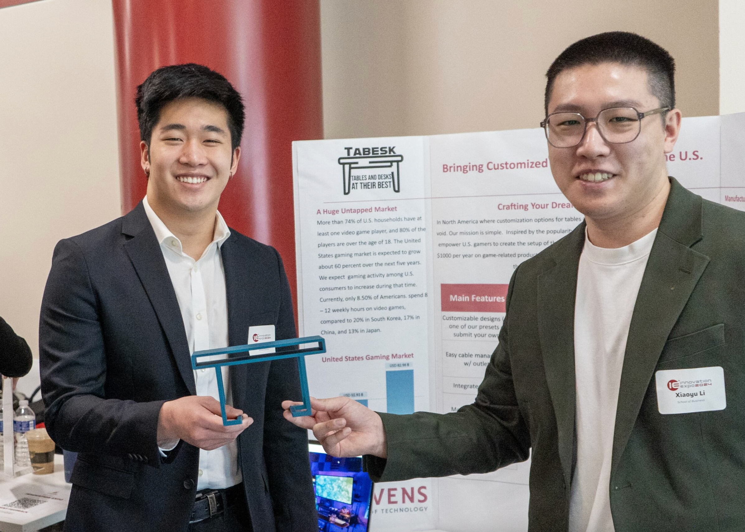 Members of the Tabesk team stand in front of their posteboard, holding a 3-D printed, small version of their product that is shaped like a table.