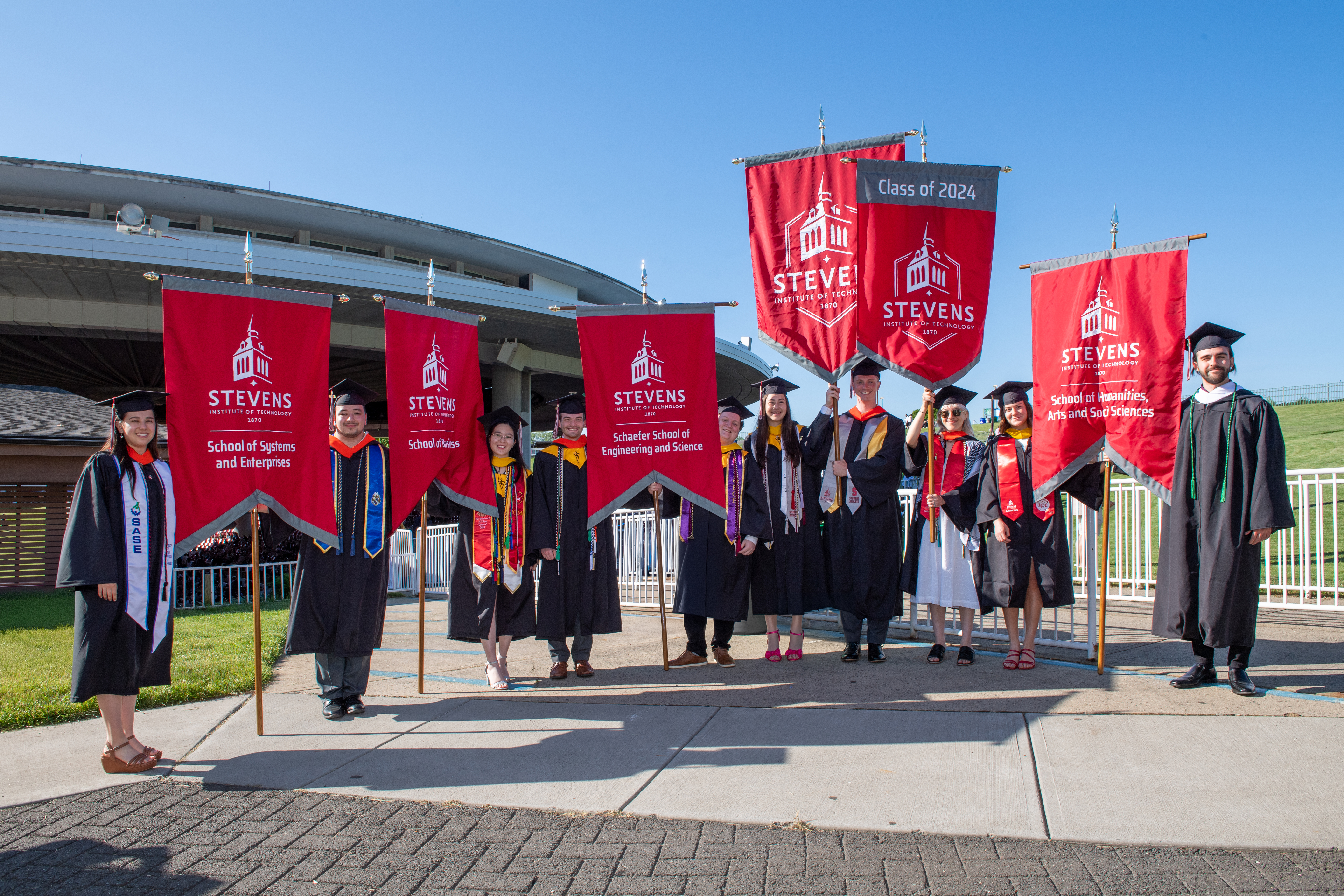 Students from each school dressed in caps and gowns hold each school's banner during Commencement 2024.