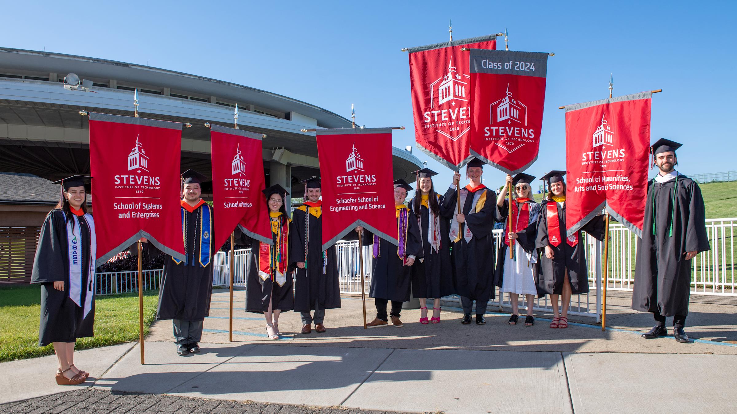 Students from each school dressed in caps and gowns hold each school's banner during Commencement 2024.