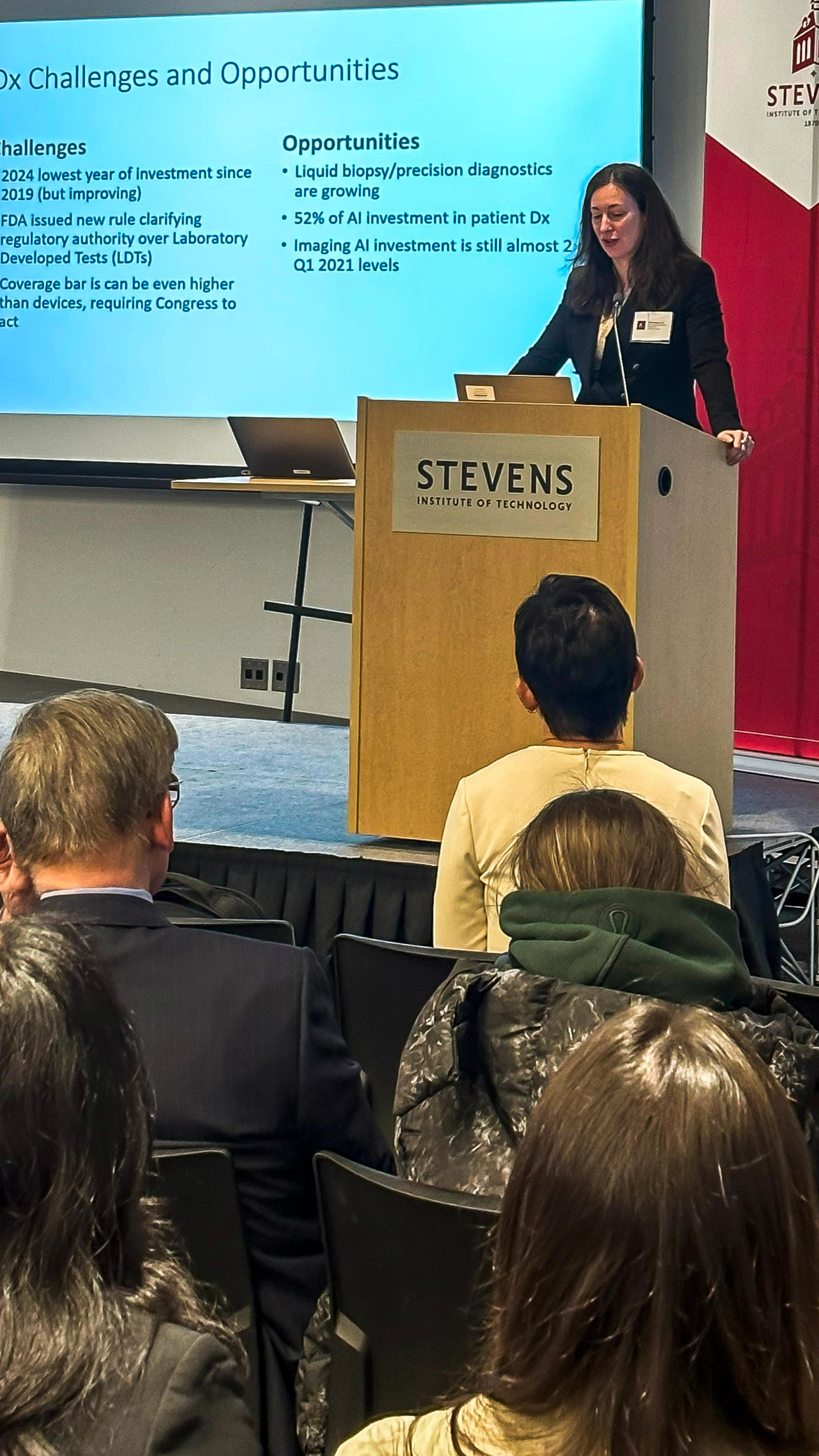 A woman in a black blouse speaks in front of a lectern with Stevens written on it, with a large-screen presentation in the background that says challenges and opportunities.