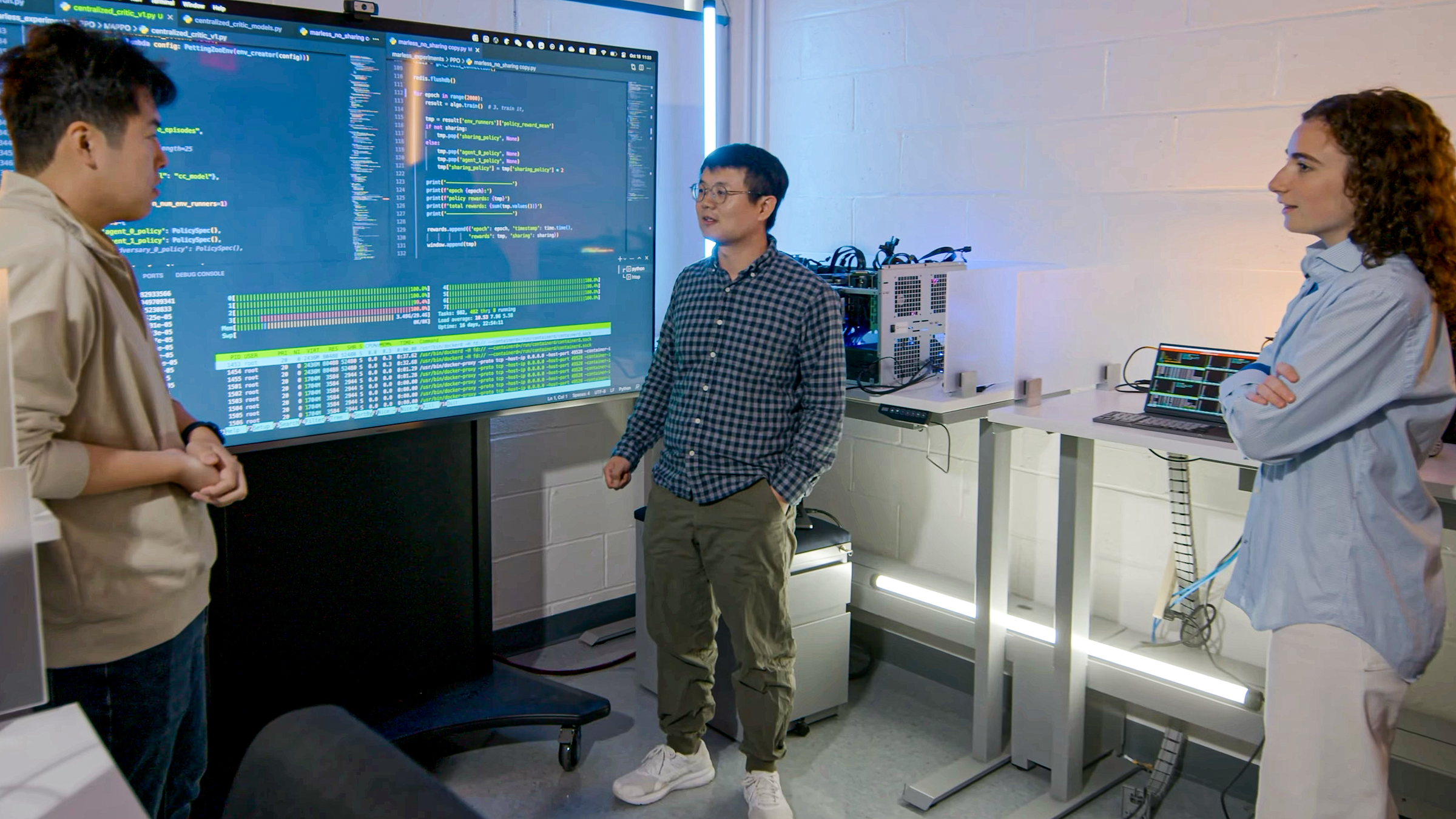Hao Wang standing between a male student on the left and a female student on the right in a lab with a big screen behind them.