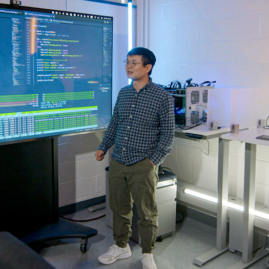 Hao Wang standing between a male student on the left and a female student on the right in a lab with a big screen behind them.