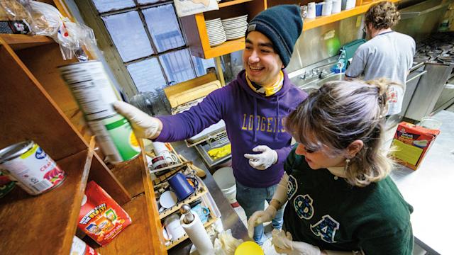A male and female student are working in a soup kitchen’s pantry. The male student is holding two cans of food that he’s reaching to place on a shelf. The female student is looking down, preparing for her next task.
