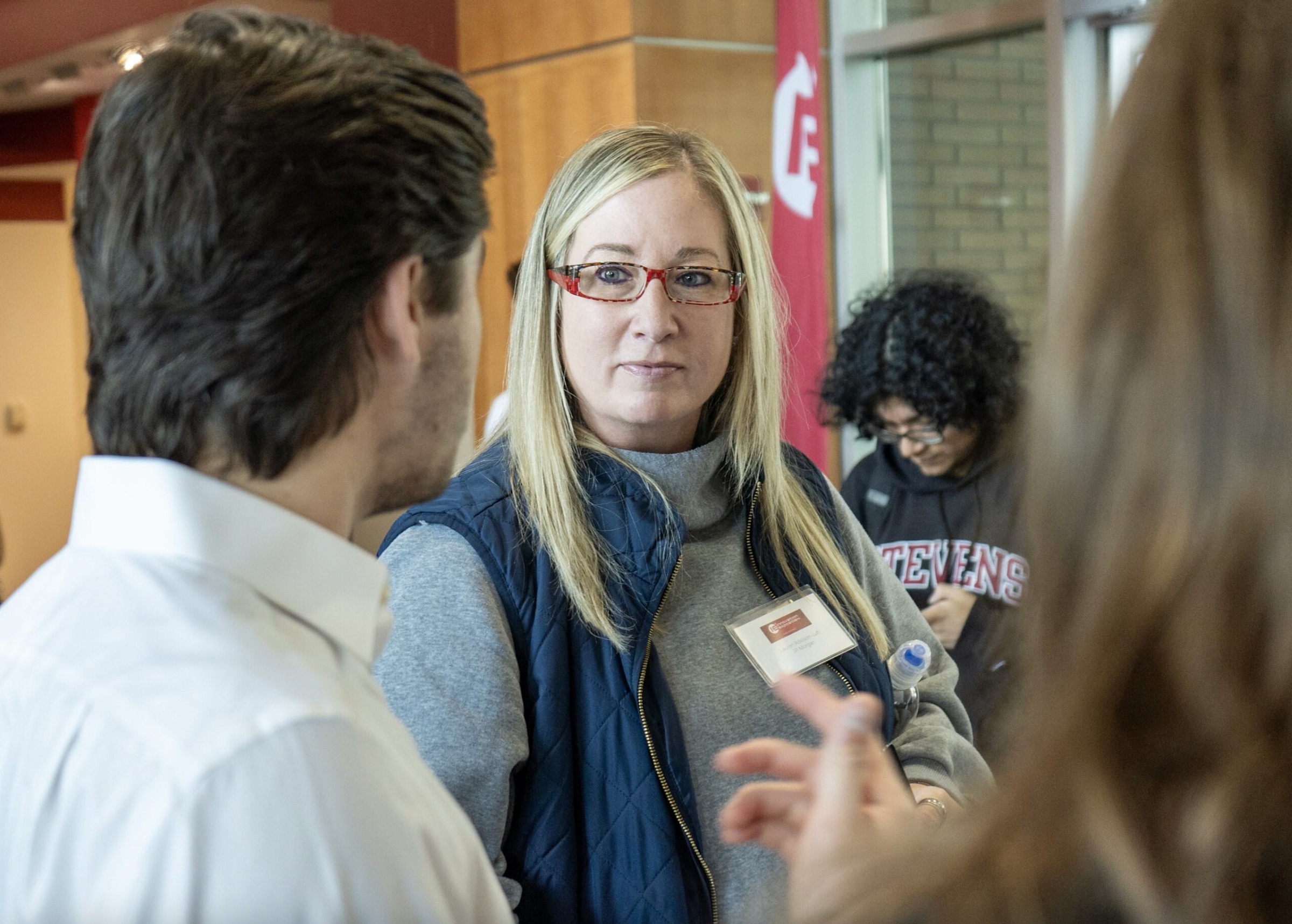 Lauren Adelson-Luft, wearing a gray sweater, blue vest and red-framed glases, speaks with students as she judges their projects.