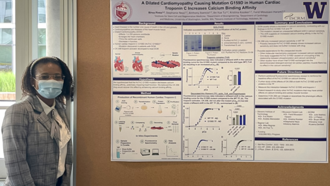 Stevens student Birva Pinto, a woman wearing a gray suit and surgeon's mask, poses next to a cork board displaying her research into cardiac disease. 